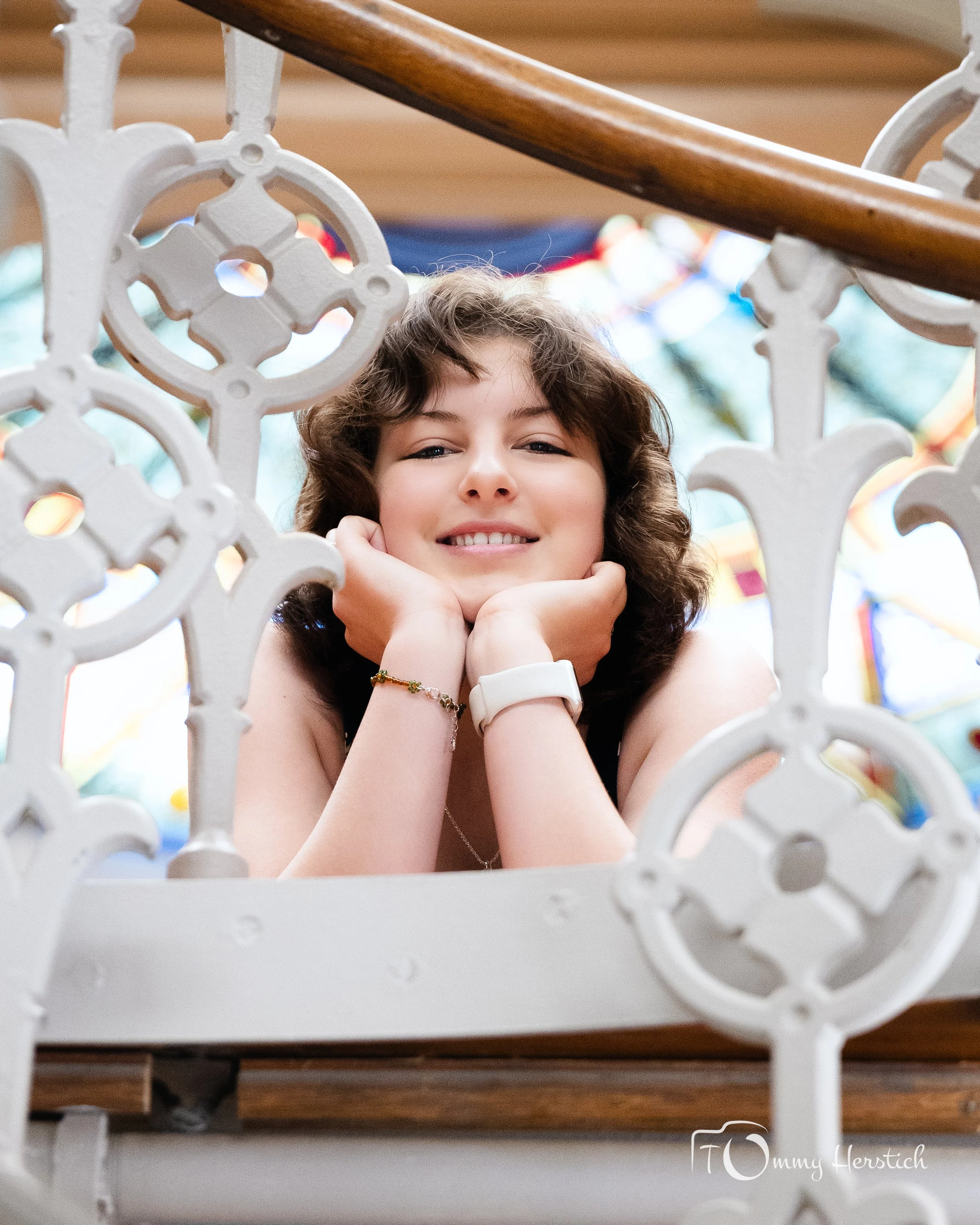 A young woman with curly hair leaning on her hands, smiling with her chin resting on her fists, seen through decorative ironwork on a staircase.