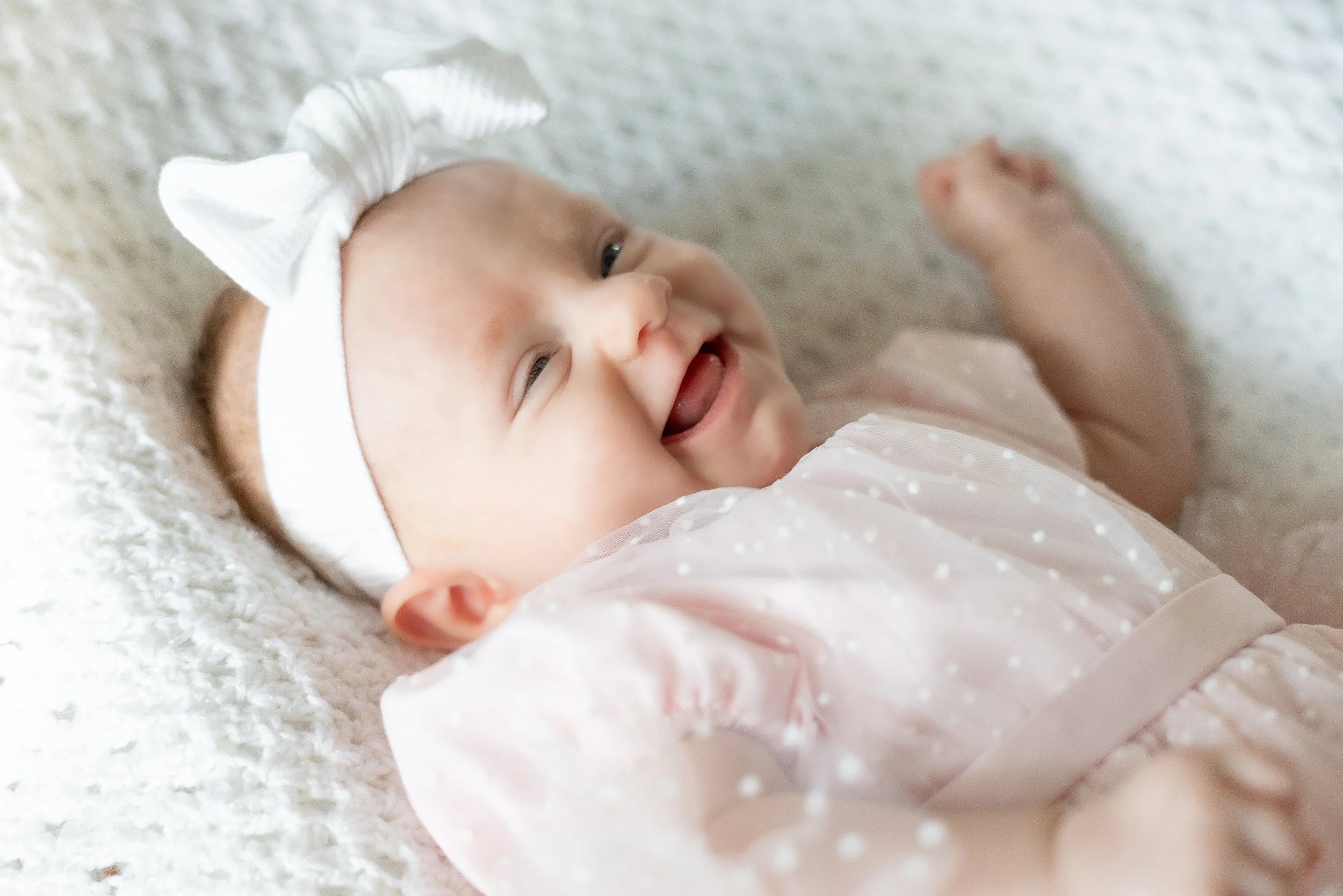 Smiling baby girl lying on a soft textured blanket, wearing a white headband with a bow and a light pink, polka-dotted dress.
