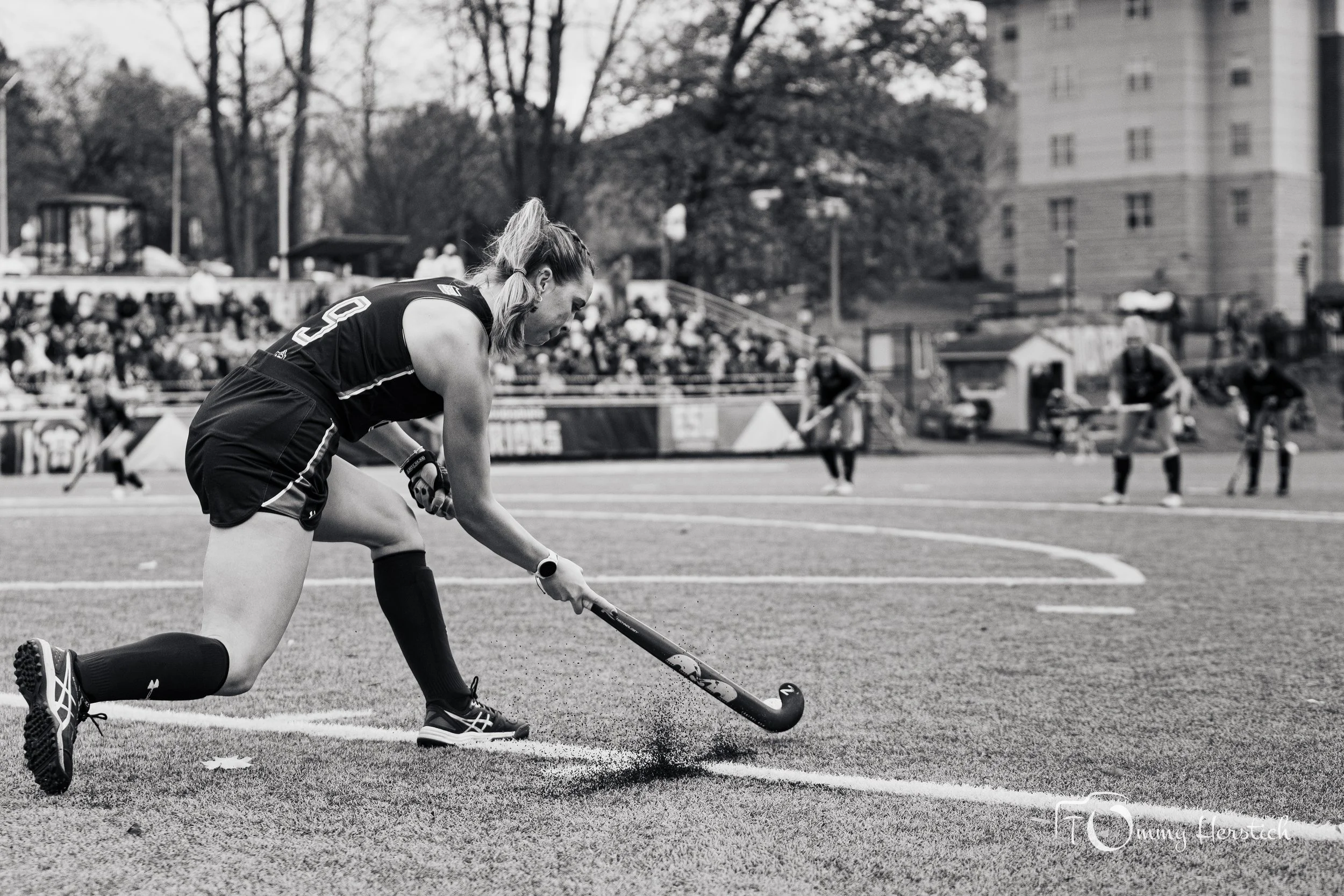 A female field hockey player in a black uniform and knee-high socks is kneeling on a field, preparing to hit a ball with her stick during a game; spectators are visible in the background.