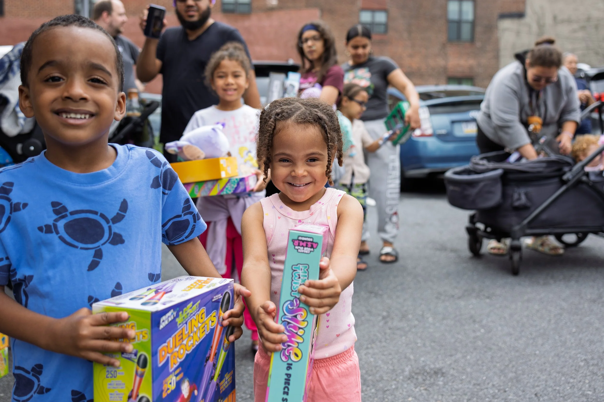 Smiling children holding toy rockets at an outdoor gathering, with adults and parked cars in the background.