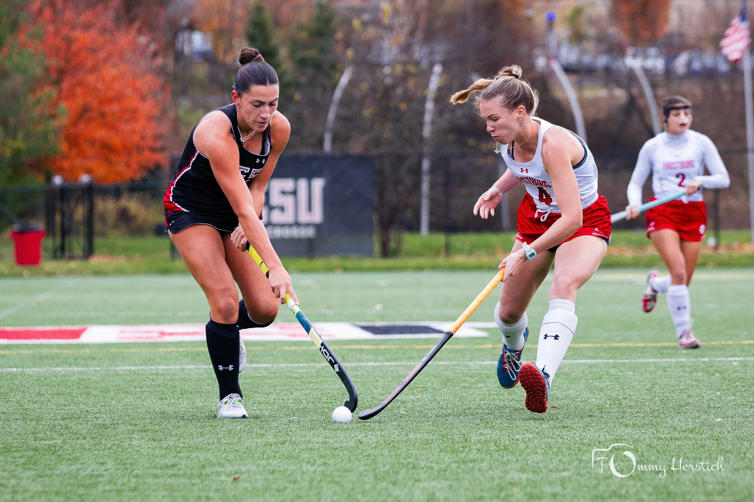 Two female field hockey players in action on a green turf field, competing for the ball. One in a black uniform and one in a white and red uniform, with another player visible in the background.