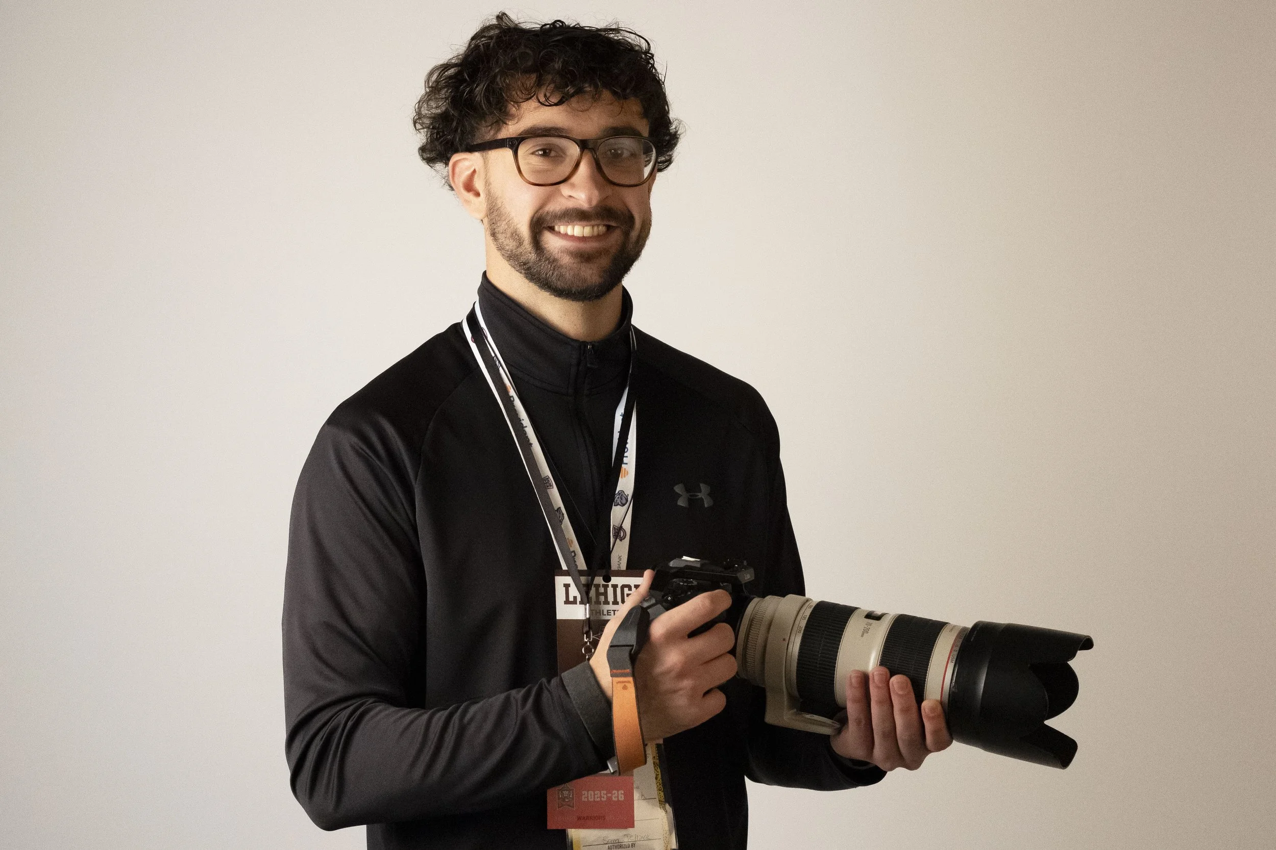 A man with curly dark hair, glasses, and a beard holding a professional camera with a large telephoto lens, smiling at the camera, wearing a black athletic jacket and two conference badges around his neck.