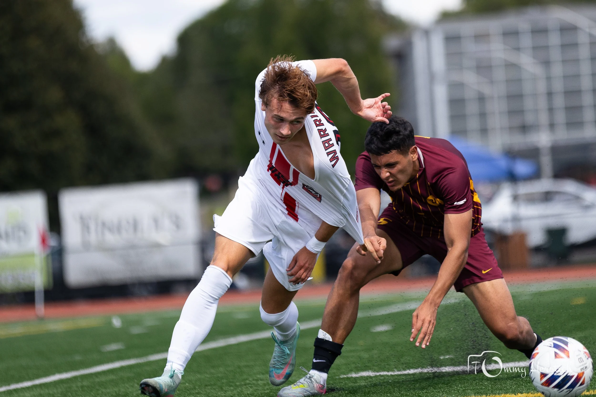 Two male soccer players competing for the ball on a field, one in a white uniform and the other in a maroon uniform, during a game.
