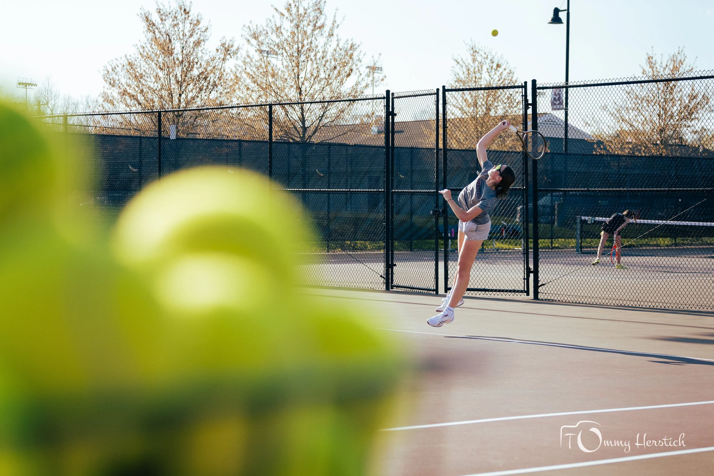 A girl in a gray t-shirt and gray shorts playing tennis on an outdoor court, about to hit a tennis ball with a tennis racket, with another person in the background also playing tennis.