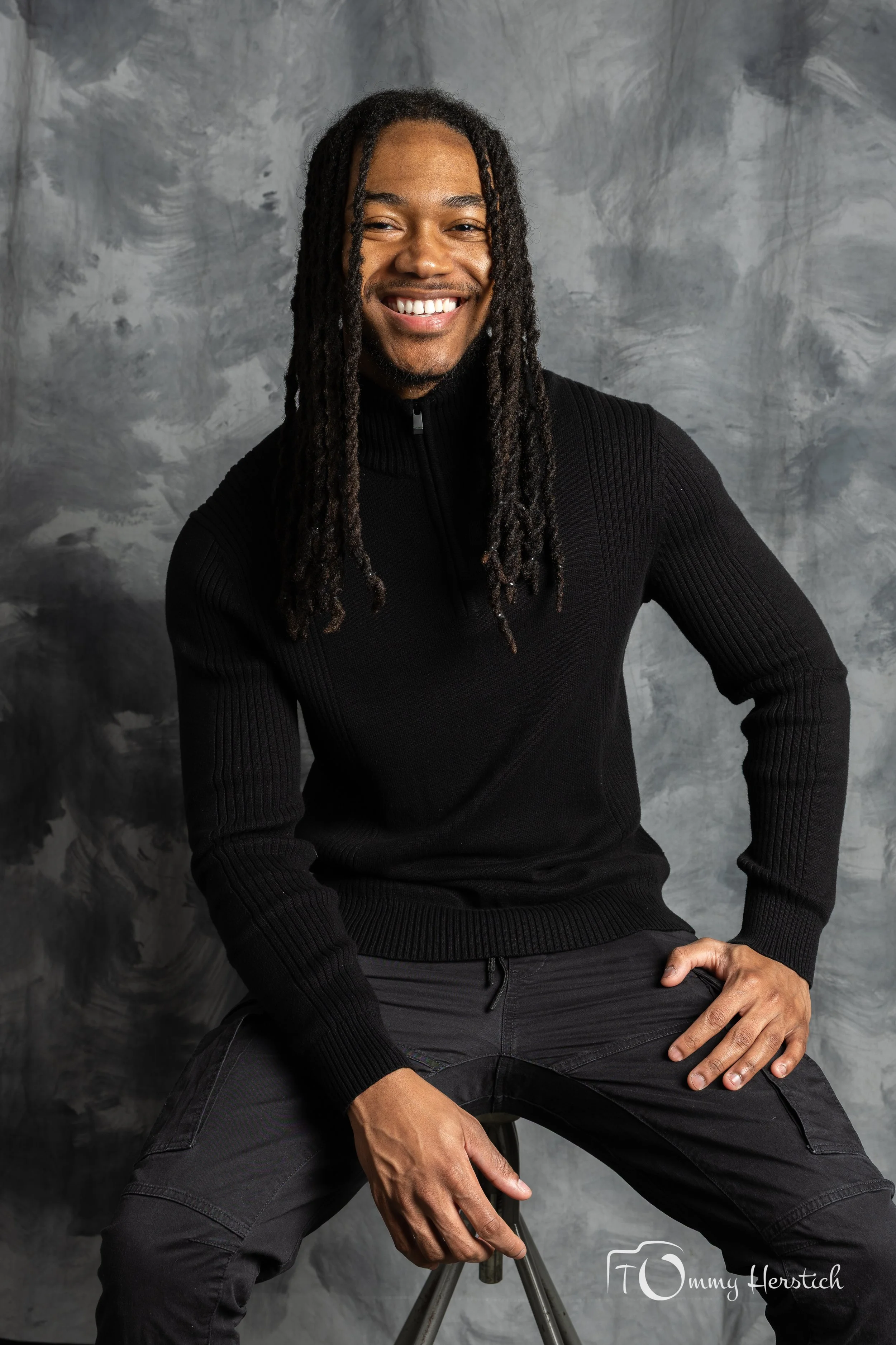 A smiling man with long dreadlocks sitting on a stool in front of a gray textured background, wearing a black sweater and black pants.