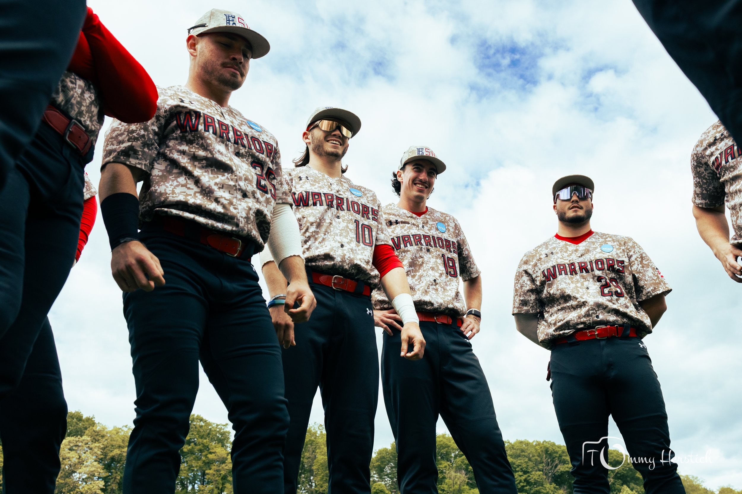 A group of five baseball players standing outdoors on a cloudy day, wearing camouflage jerseys with 'WARRIORS' and numbers, dark pants, and some wearing sunglasses or caps, appearing confident.