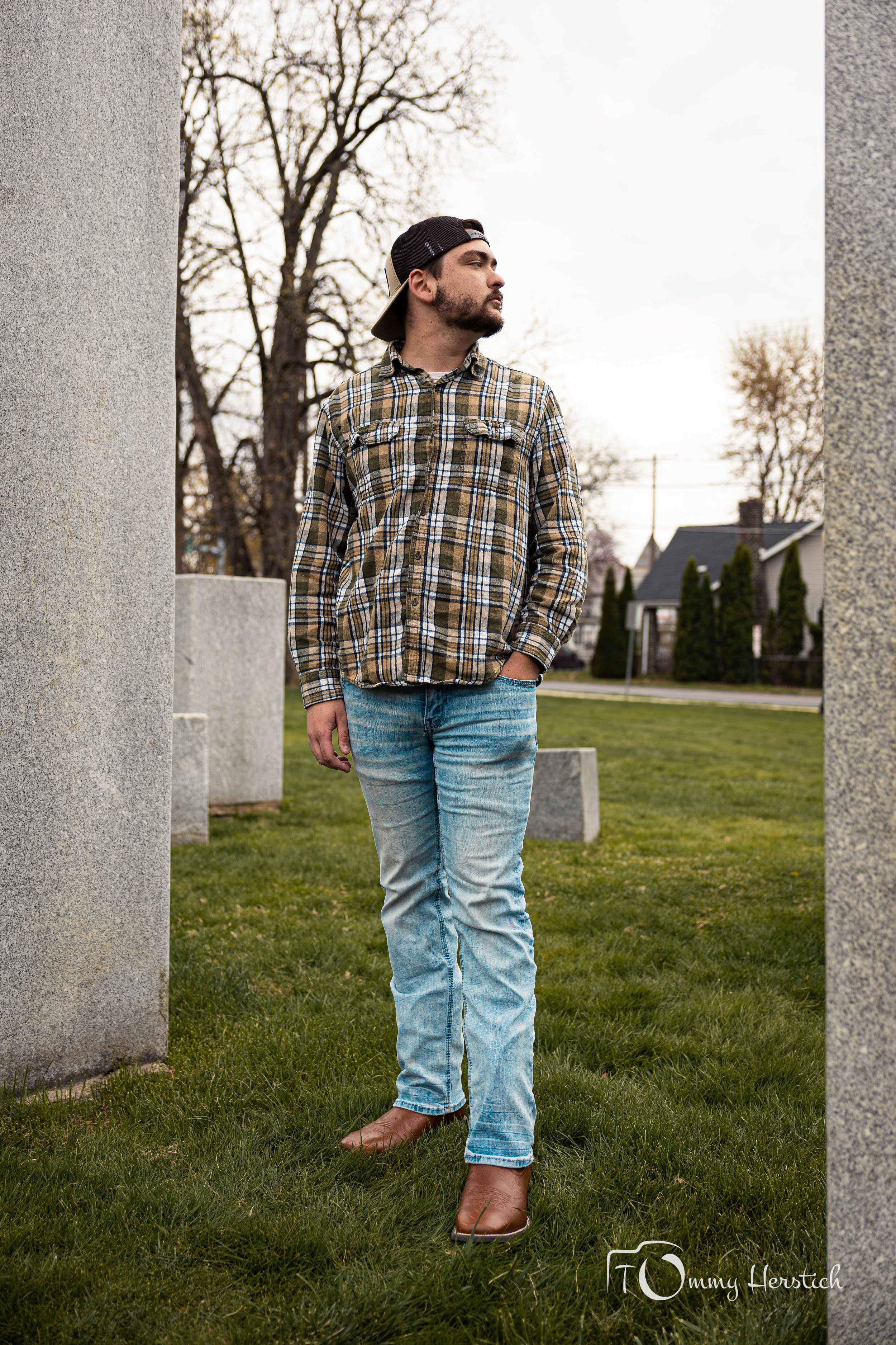 A young man in a plaid shirt and blue jeans walking through a park surrounded by large stone monuments, with trees and houses in the background.