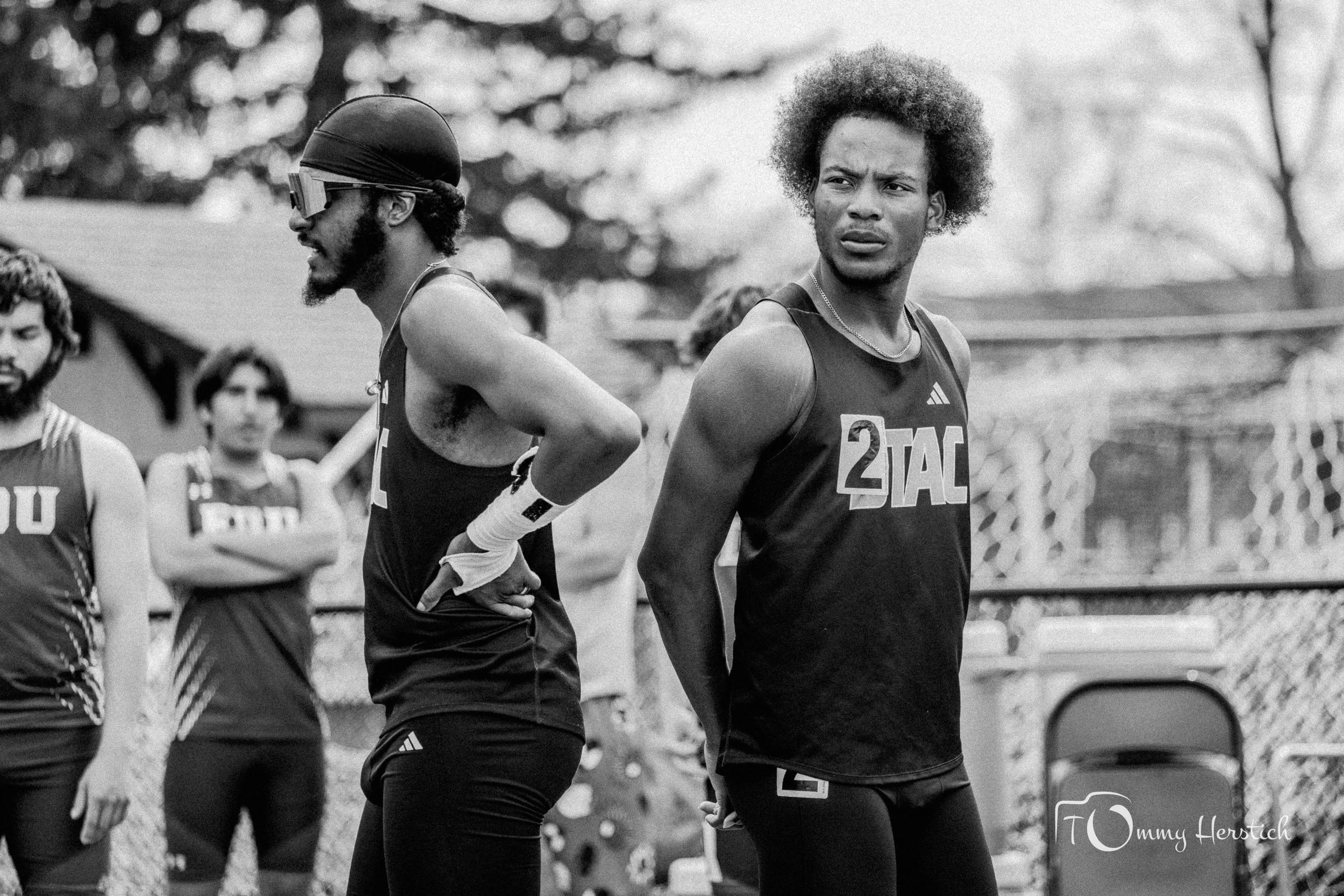 Black-and-white photo of male athletes at a track and field event, wearing athletic uniforms and standing outdoors. One athlete is in the foreground, slightly looking to the side, with others in the background.