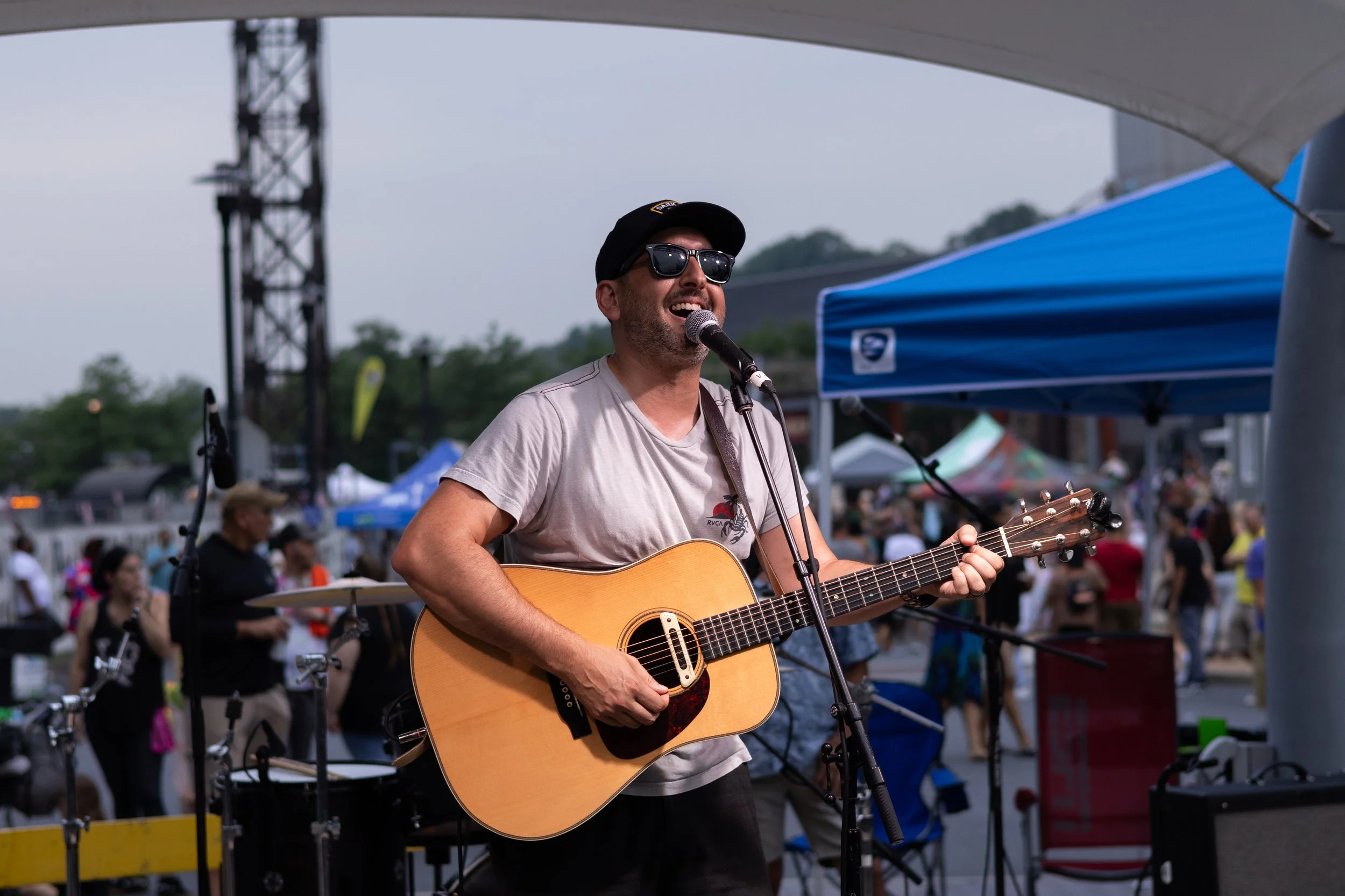 A man wearing sunglasses, a cap, and a T-shirt is singing and playing an acoustic guitar at an outdoor event, with a crowd and tents in the background.