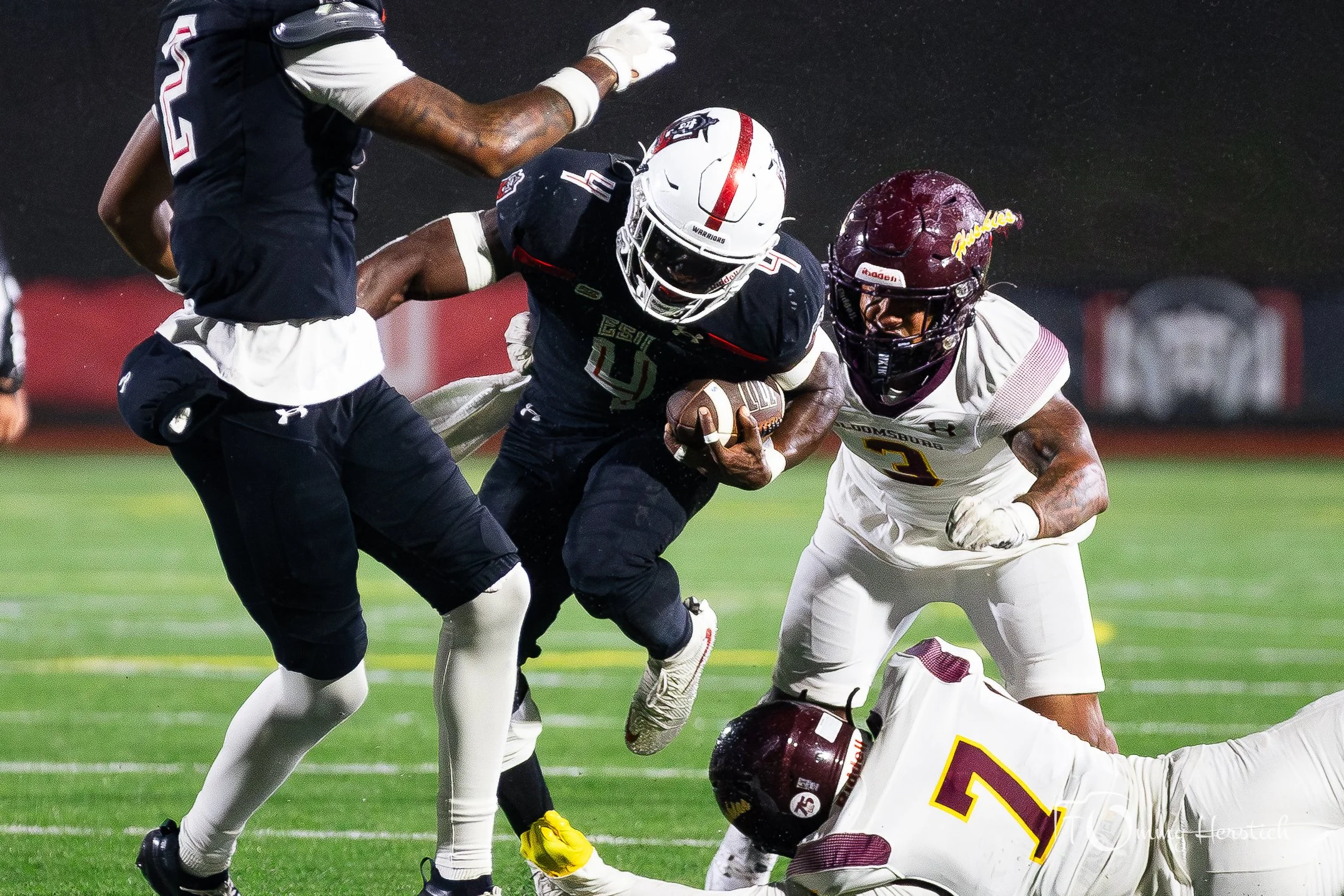 American football game with players: one in a dark uniform carrying the ball, others in white uniforms trying to tackle him on a field at night.