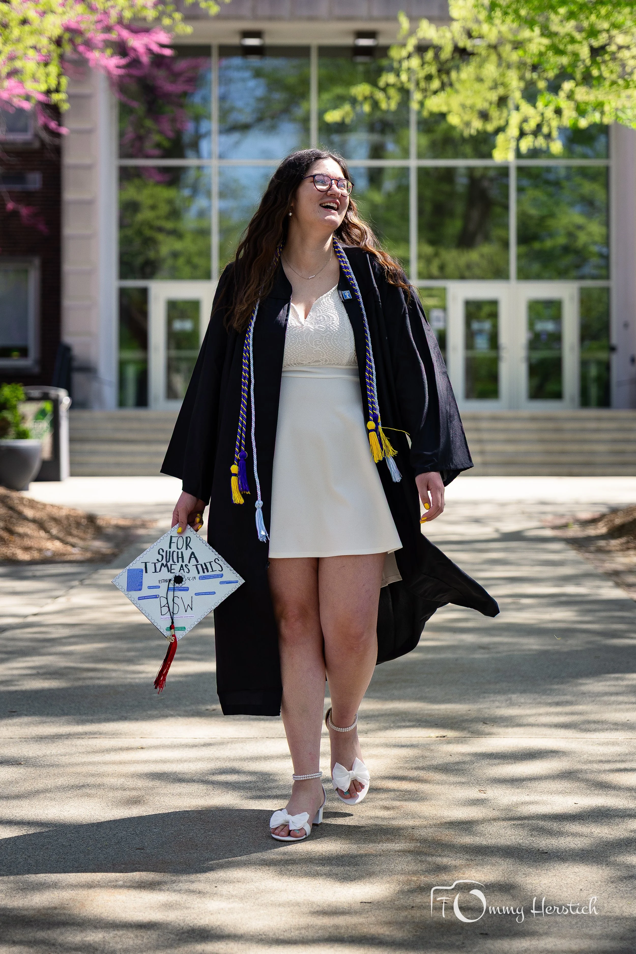 A woman in a graduation cap and gown walking outdoors in front of a modern building with glass windows. She is smiling, holding her decorated graduation cap, and wearing white high heels. The background has trees and sunlight.