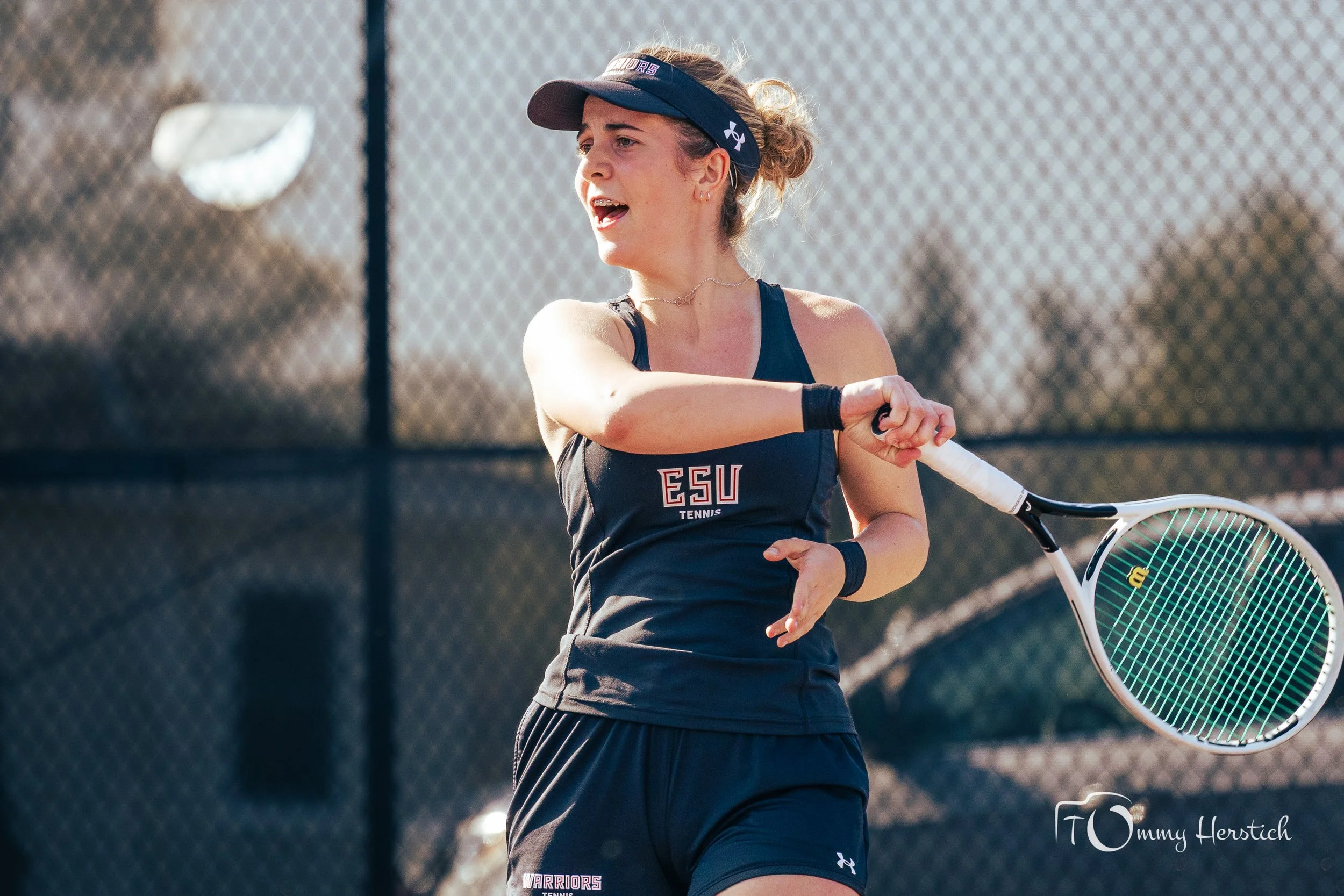 A female tennis player wearing a black sports uniform with 'ESU Tennis' written on it, a black visor, and black wristbands, preparing to hit a tennis ball with her racket on an outdoor court.