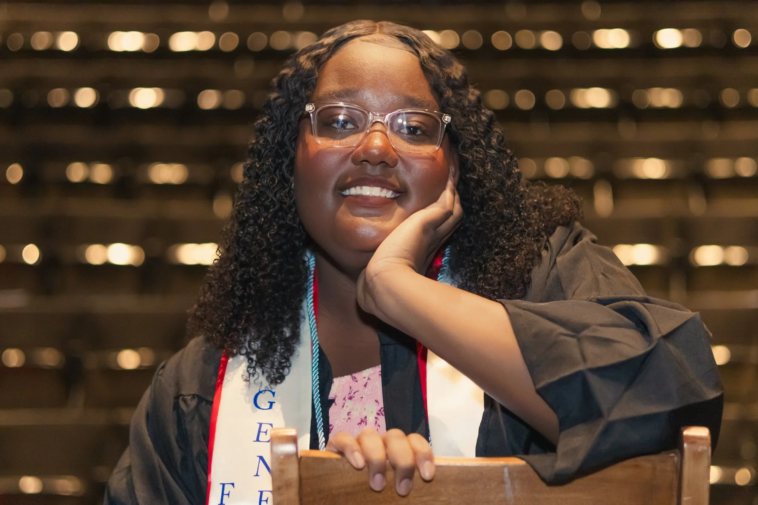 A woman with glasses, curly hair, and a graduation gown sitting in a auditorium, smiling and resting her chin on her hand.