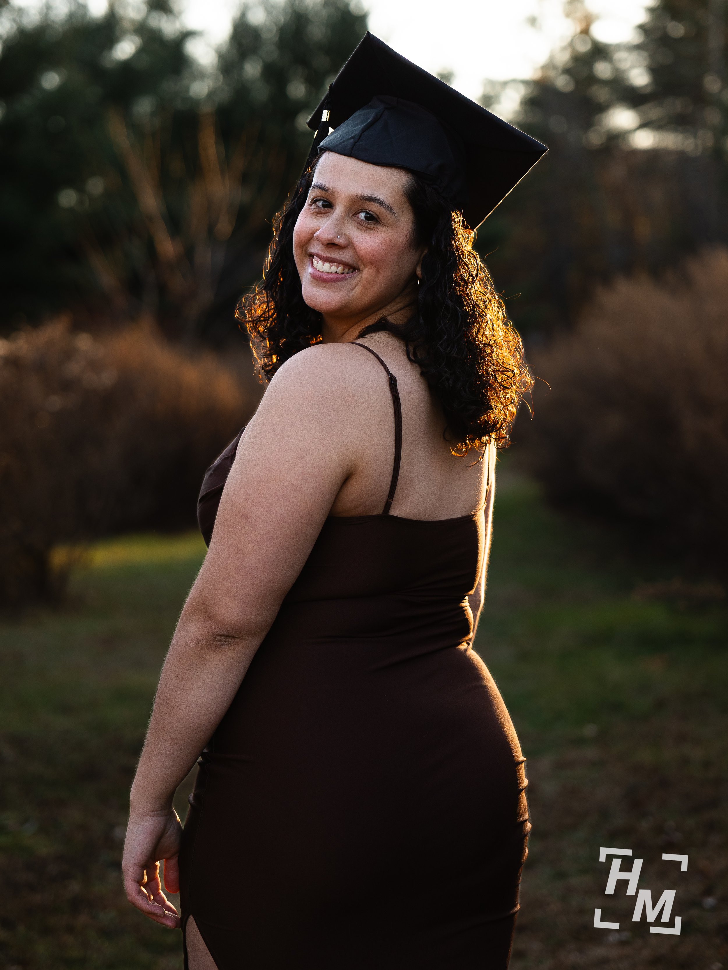 A woman smiling while wearing a graduation cap and dress outdoors during sunset.