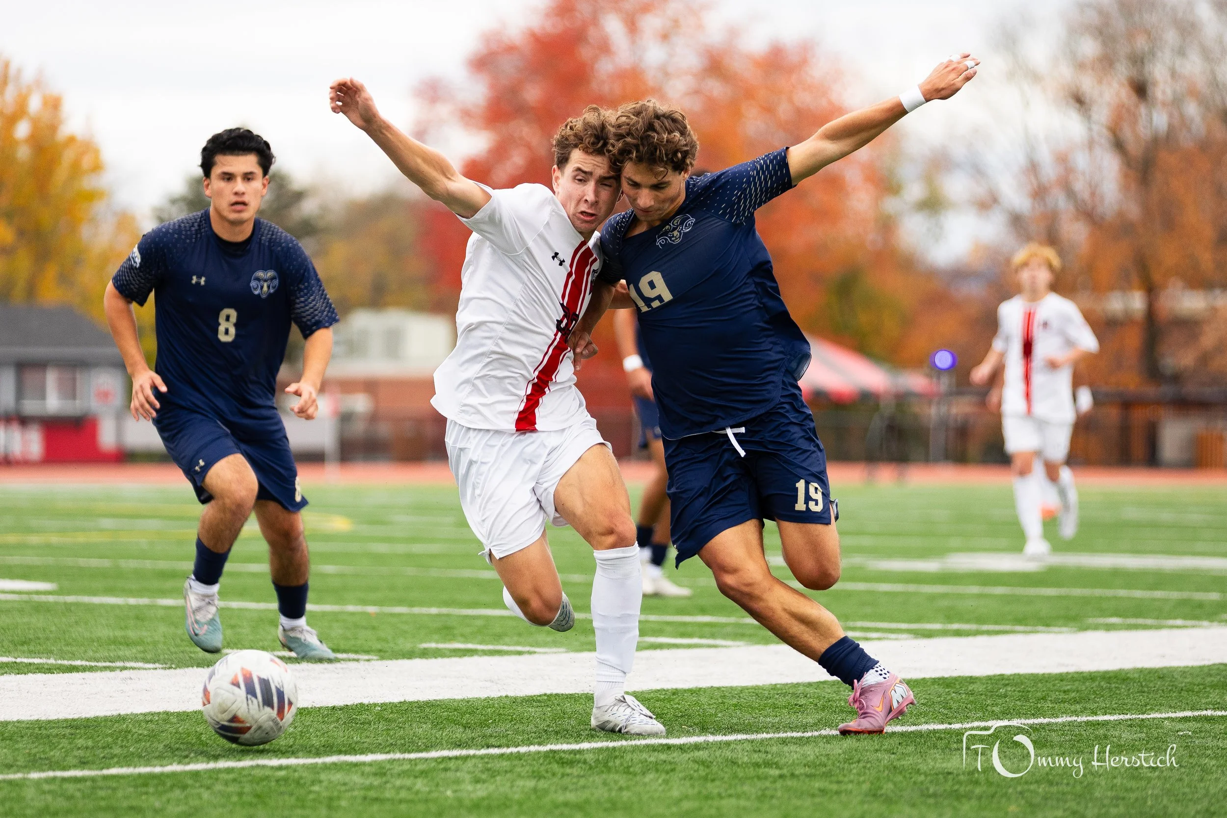 Two soccer players are competing for the ball on a soccer field during daytime, with other players and trees with autumn foliage in the background.