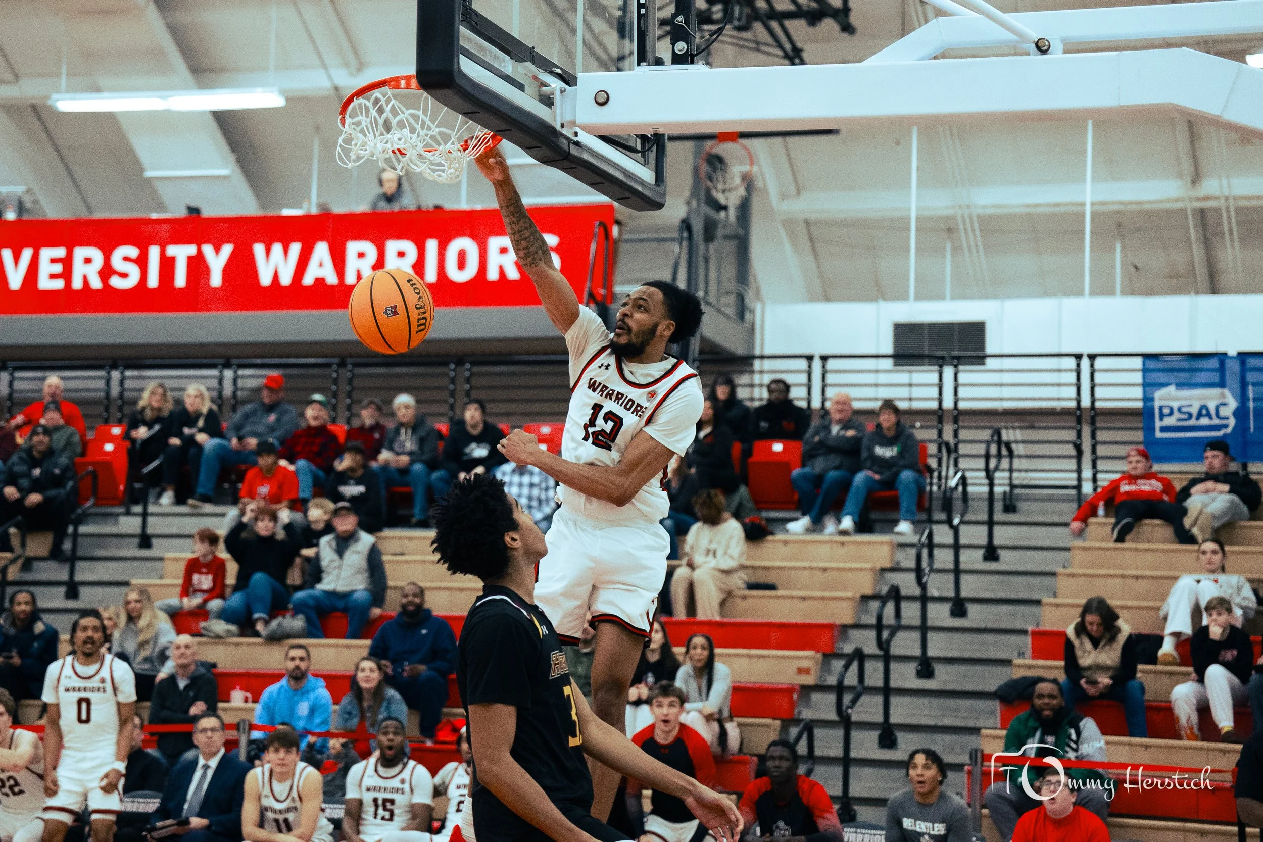 A basketball player with jersey number 12 jumps for a dunk in a gymnasium during a game, with an audience seated on bleachers in the background.