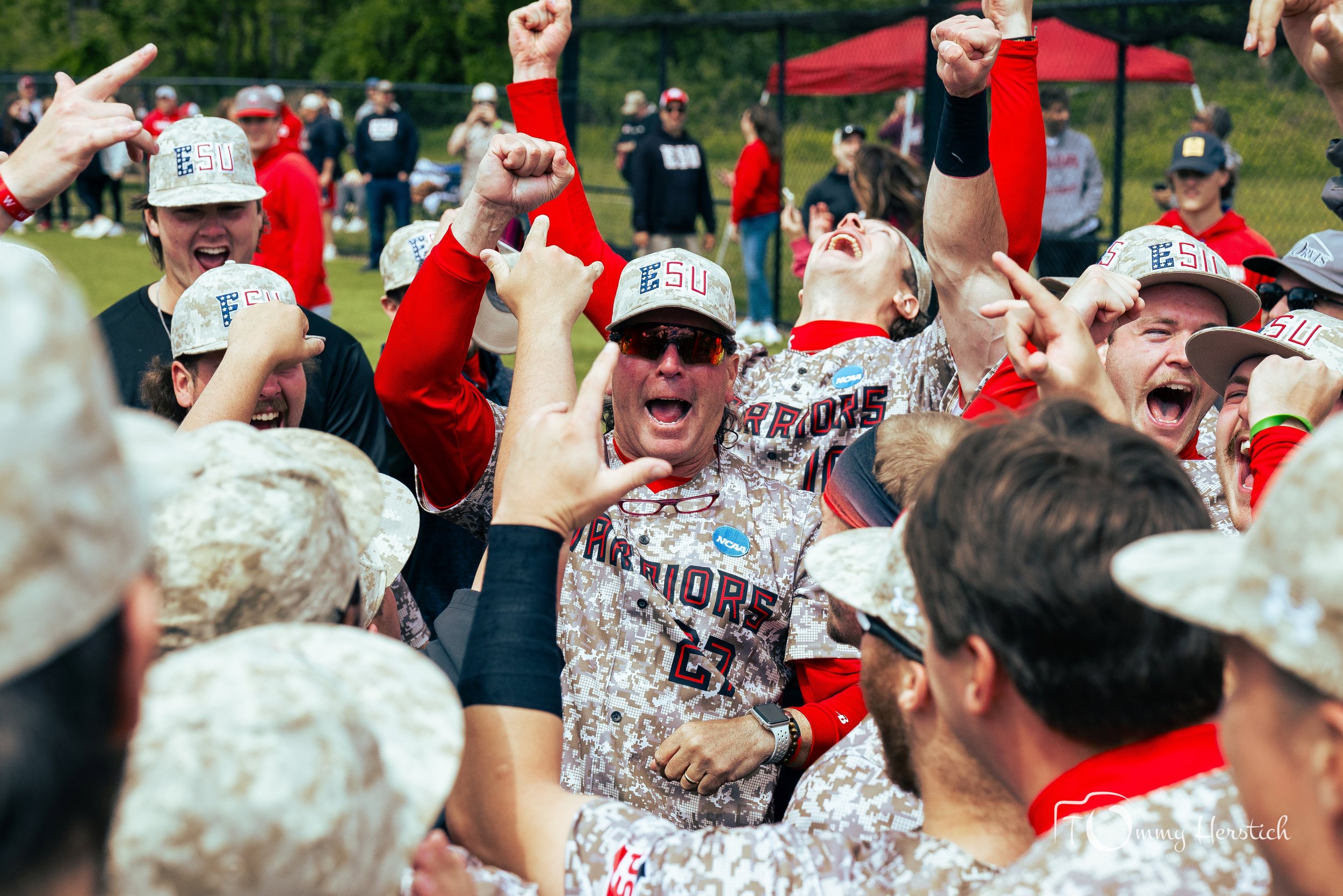 A group of people celebrating, wearing camouflage jerseys and hats with 'ESU' and 'Warriors' on them, outdoors on a sports field.