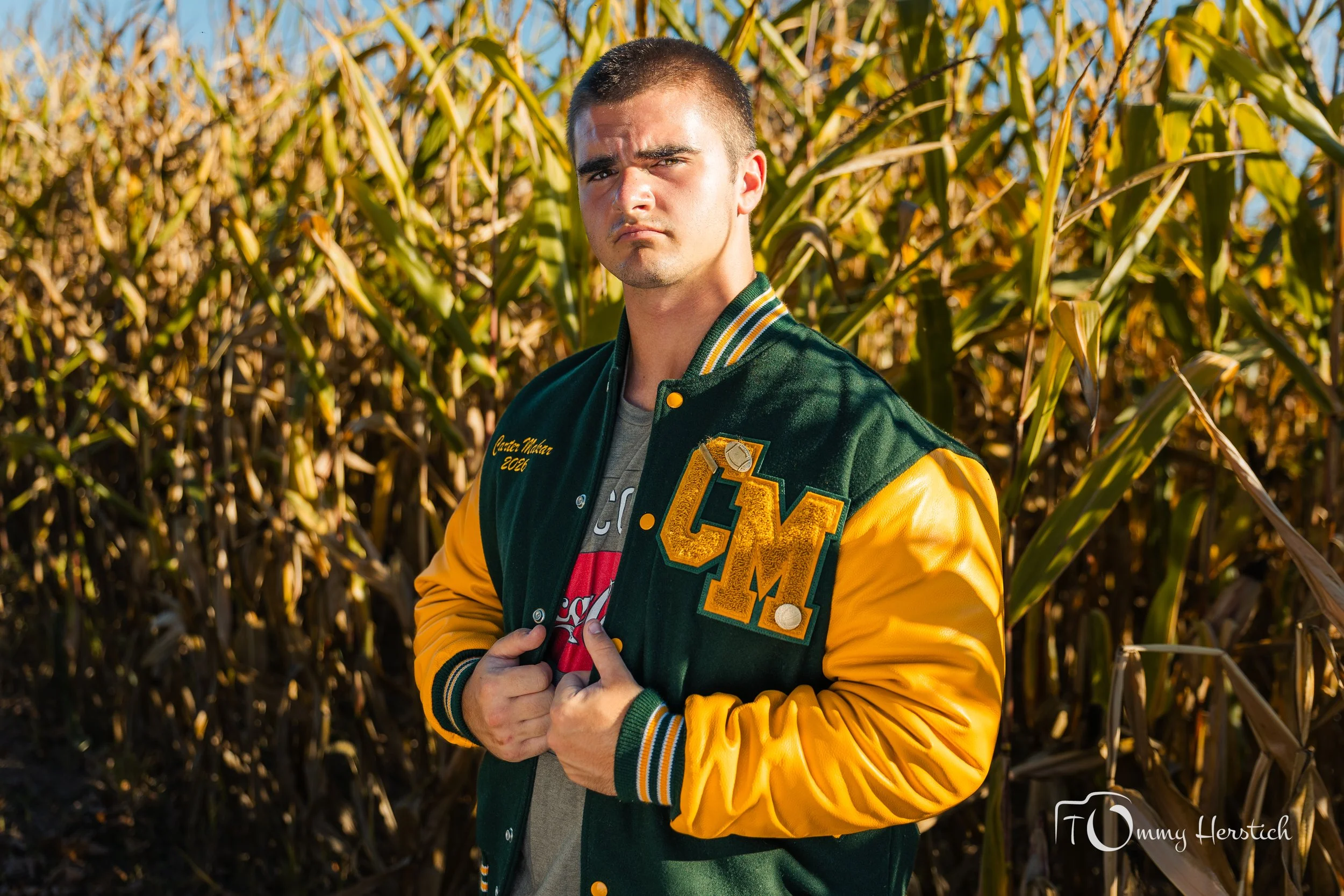A young man standing in a cornfield wearing a green and yellow varsity jacket with large embroidered letters and patches, a gray t-shirt underneath, and holding the front of his jacket with both hands.