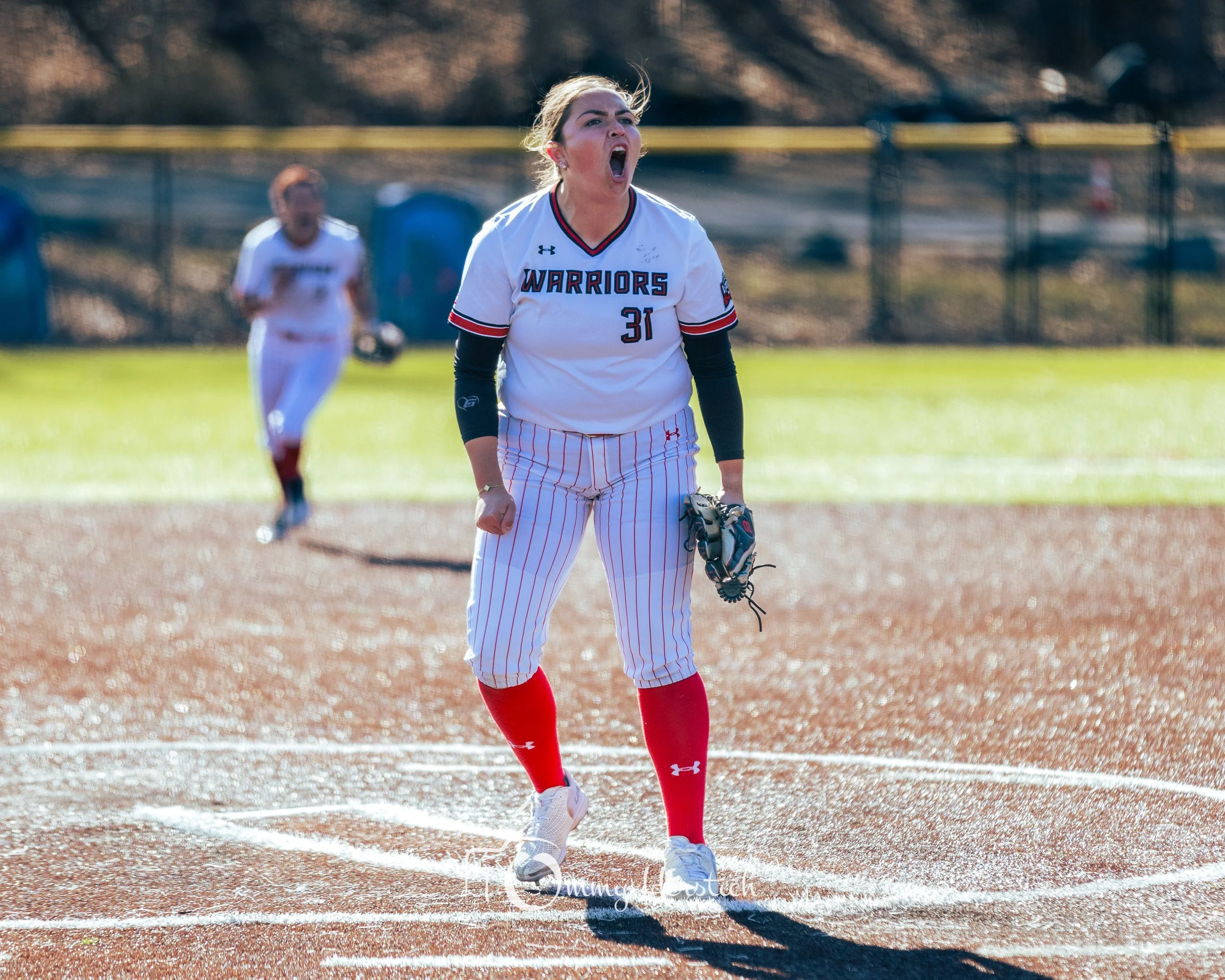 A female softball player in a Warriors uniform, wearing the number 31, shouting on the field with a glove in her left hand, with another player in the background.