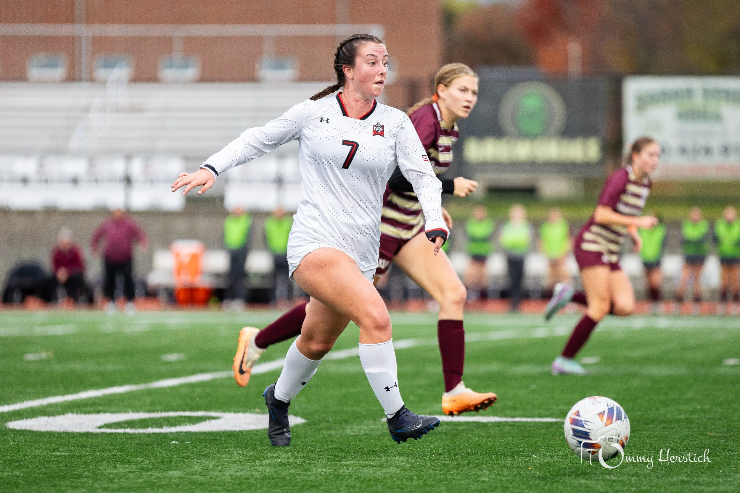 A female soccer player in a white jersey numbered 7 dribbling a soccer ball on a green field, with two players in maroon and gold striped jerseys running in the background.
