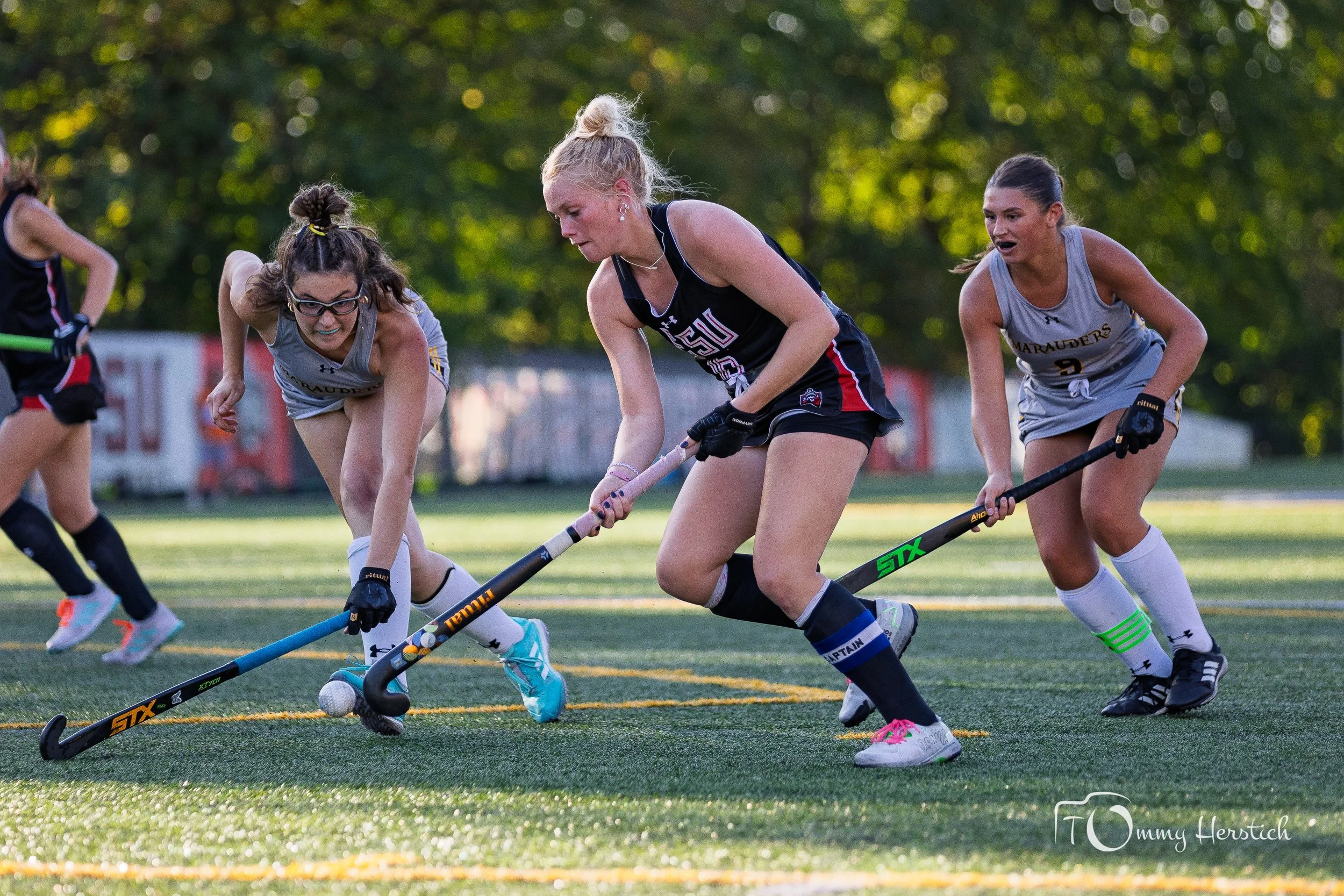 Three female field hockey players competing for the ball on a grass field during a game, with trees in the background.