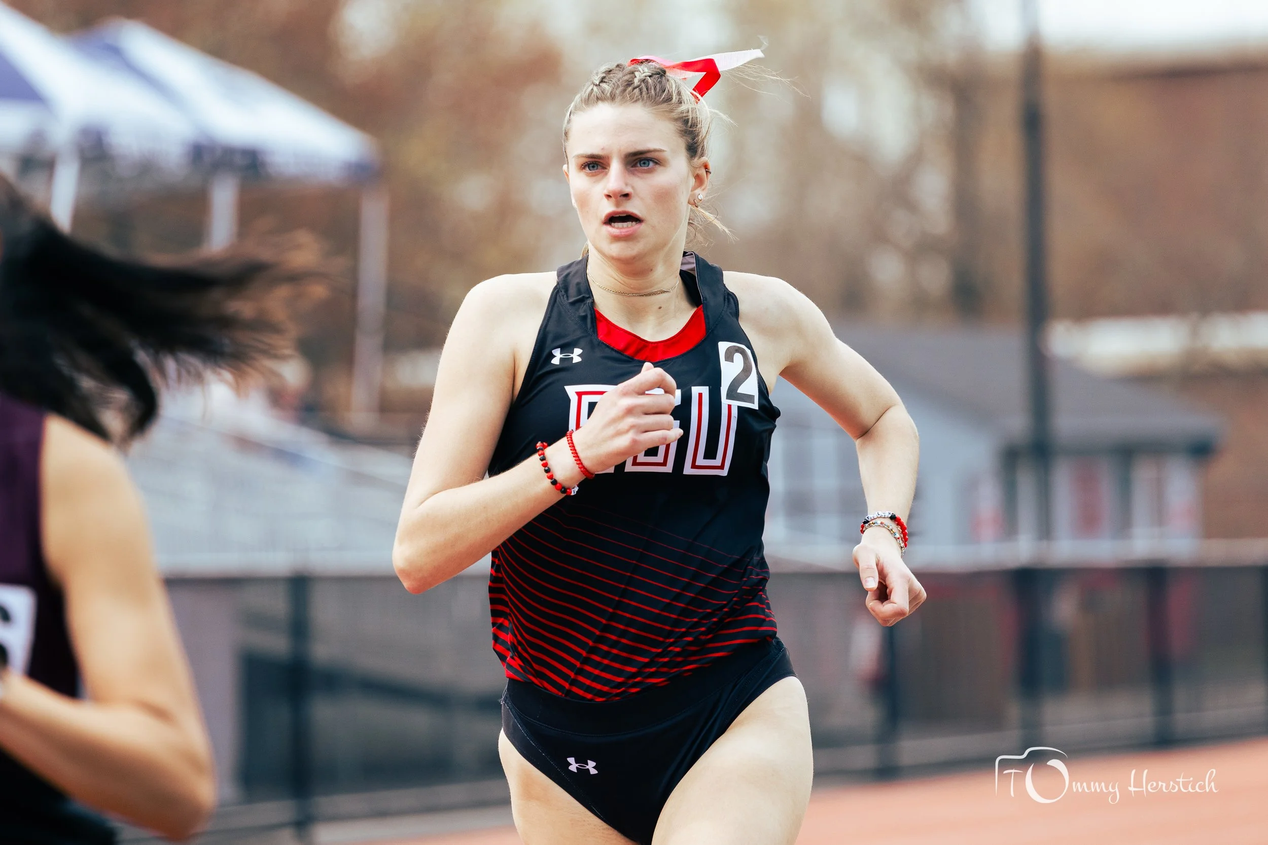 A young female athlete running on an outdoor track during a race. She has light hair tied back with a red and white ribbon, wearing a black and red running jersey and black shorts. She is looking focused and determined.