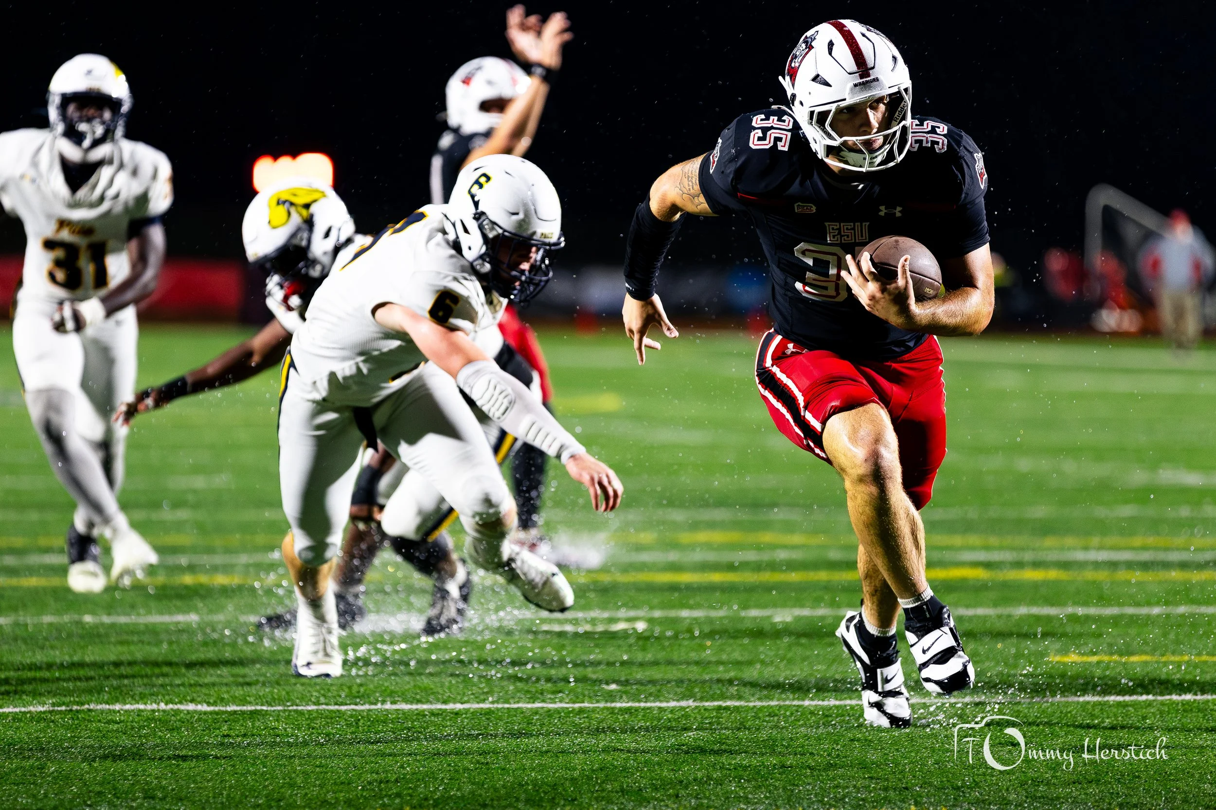 A football player in a black and red uniform running with the ball while being chased by players in white uniforms during a night game.