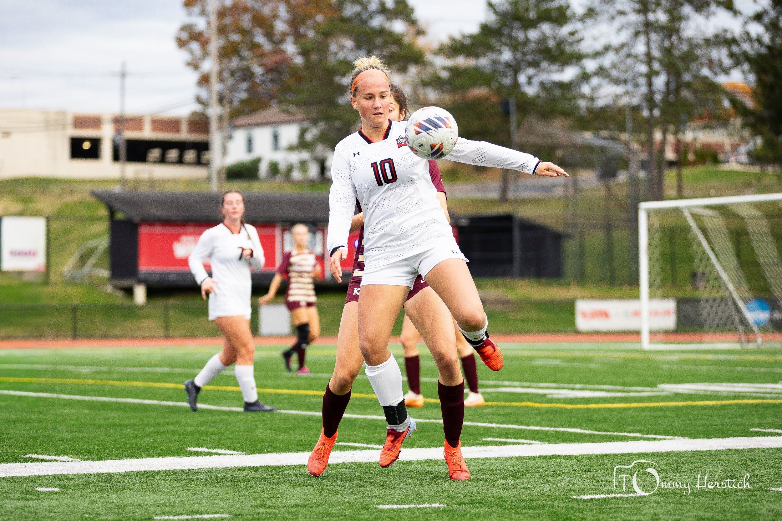 A female soccer player in a white uniform, number 10, jumping to control the ball during a game on a green field. Other players in uniforms are visible in the background.