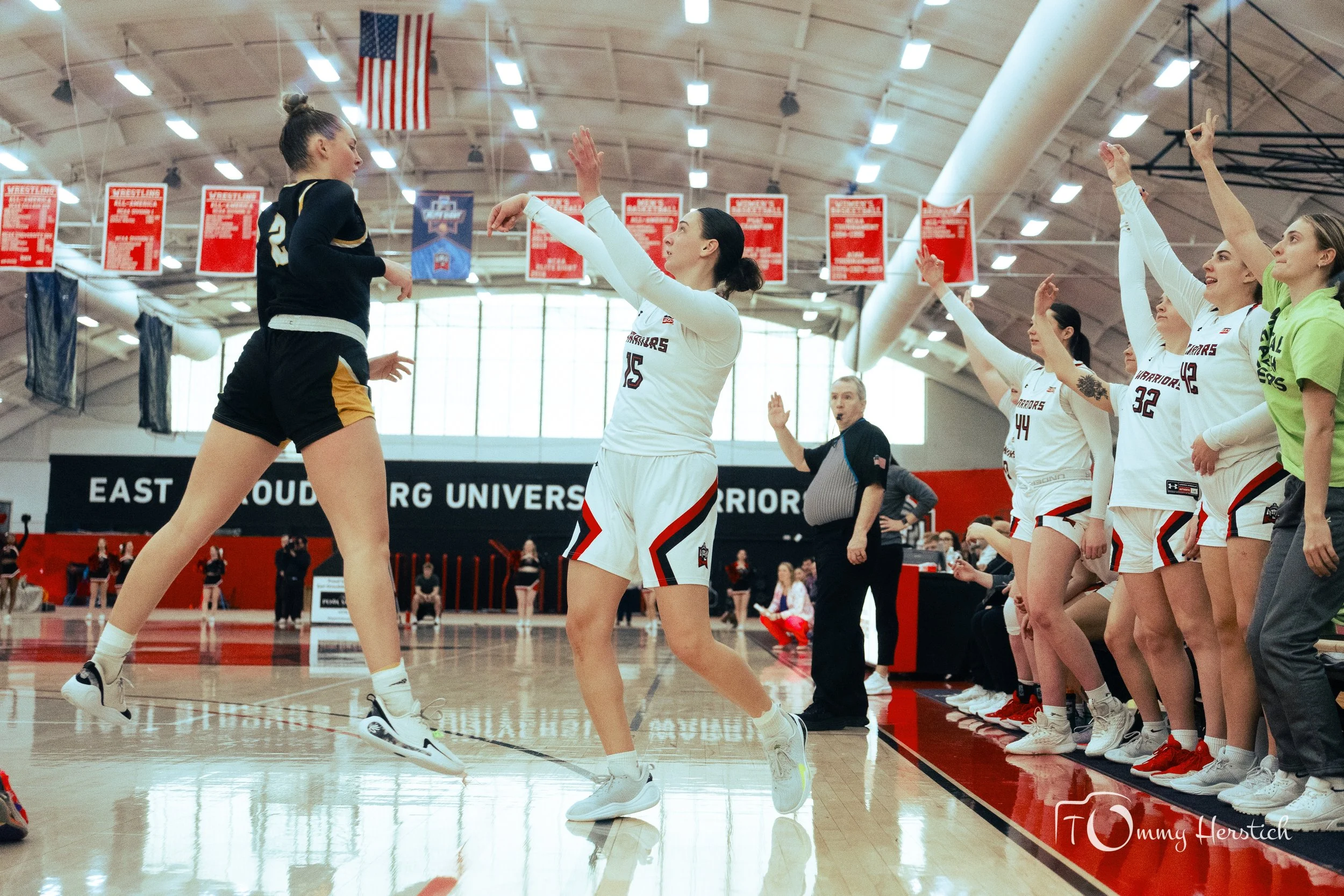 A women's basketball game action shot with players and coach, team in white jerseys, and a girl in black jersey jumping on the court, in an indoor gym with banners and an American flag hanging from the ceiling.