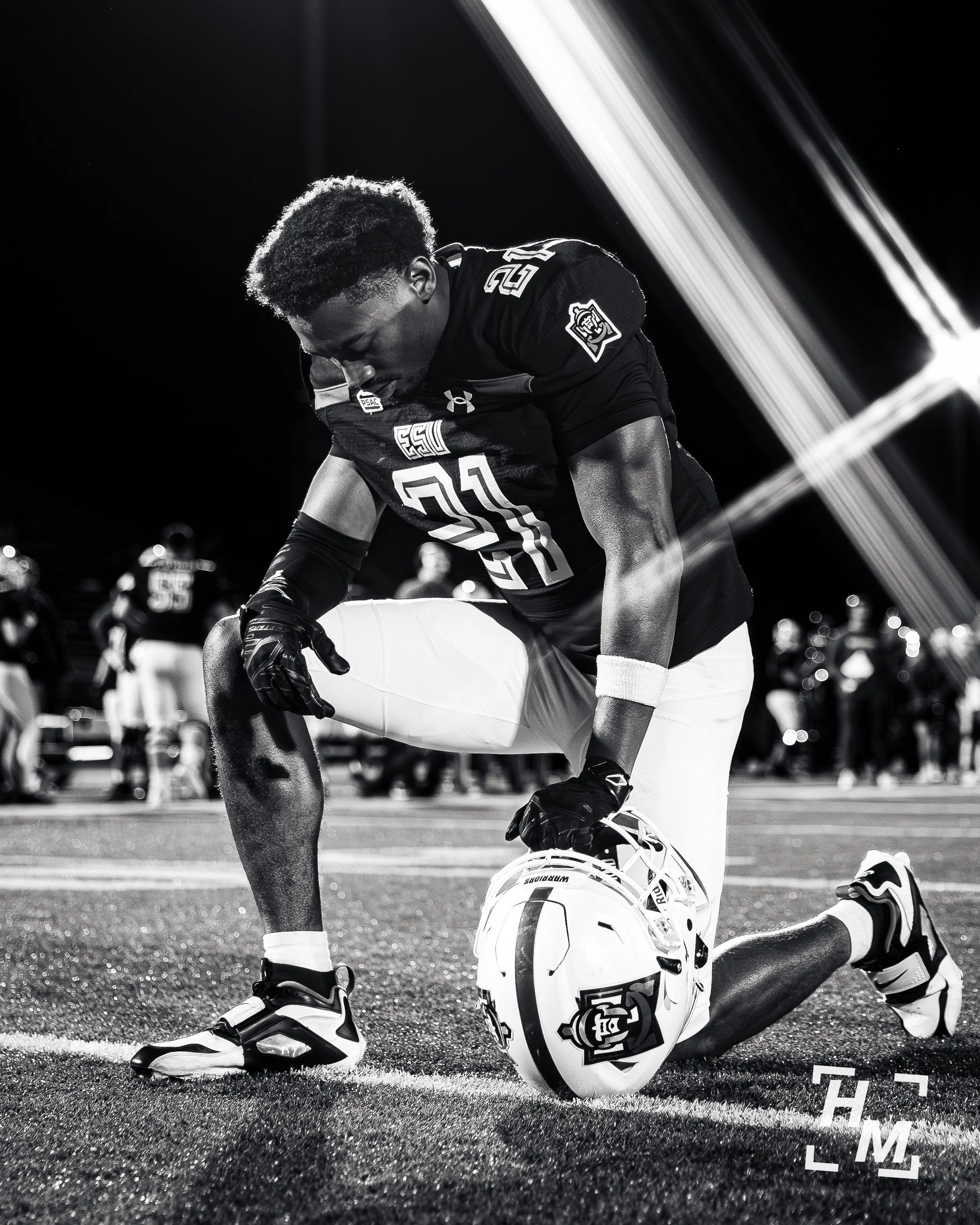 A football player kneeling on the field, holding his helmet, with his head bowed in prayer at night, surrounded by other players and spectators.