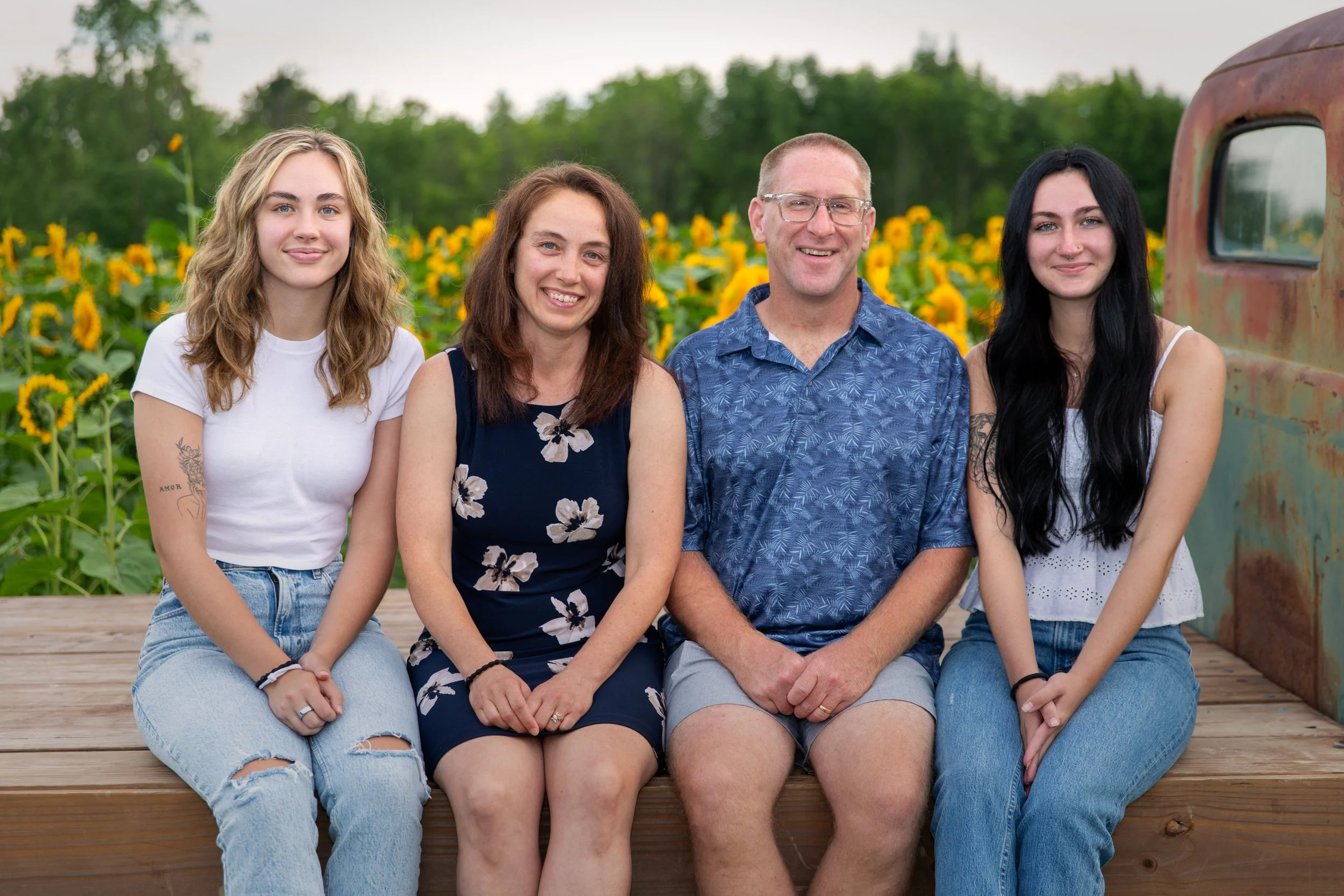 A family of four sitting on a wooden platform outdoors during daytime, with a sunflower field and green trees in the background.