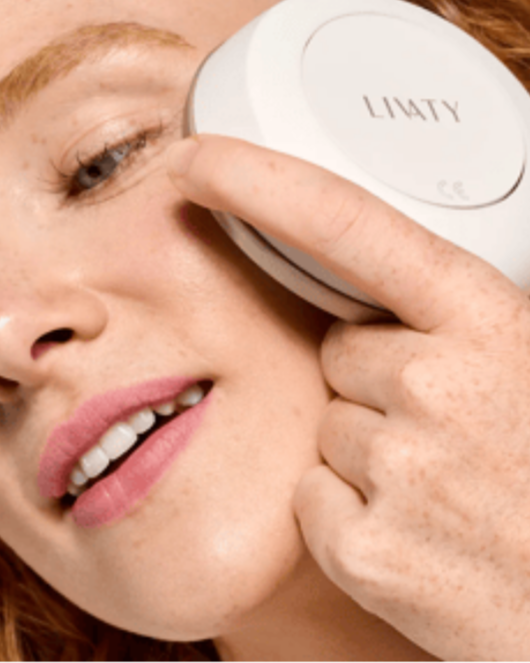 A woman with light skin and freckles is holding a LITTY beauty device near her eye, with her eyes closed and lips slightly parted.