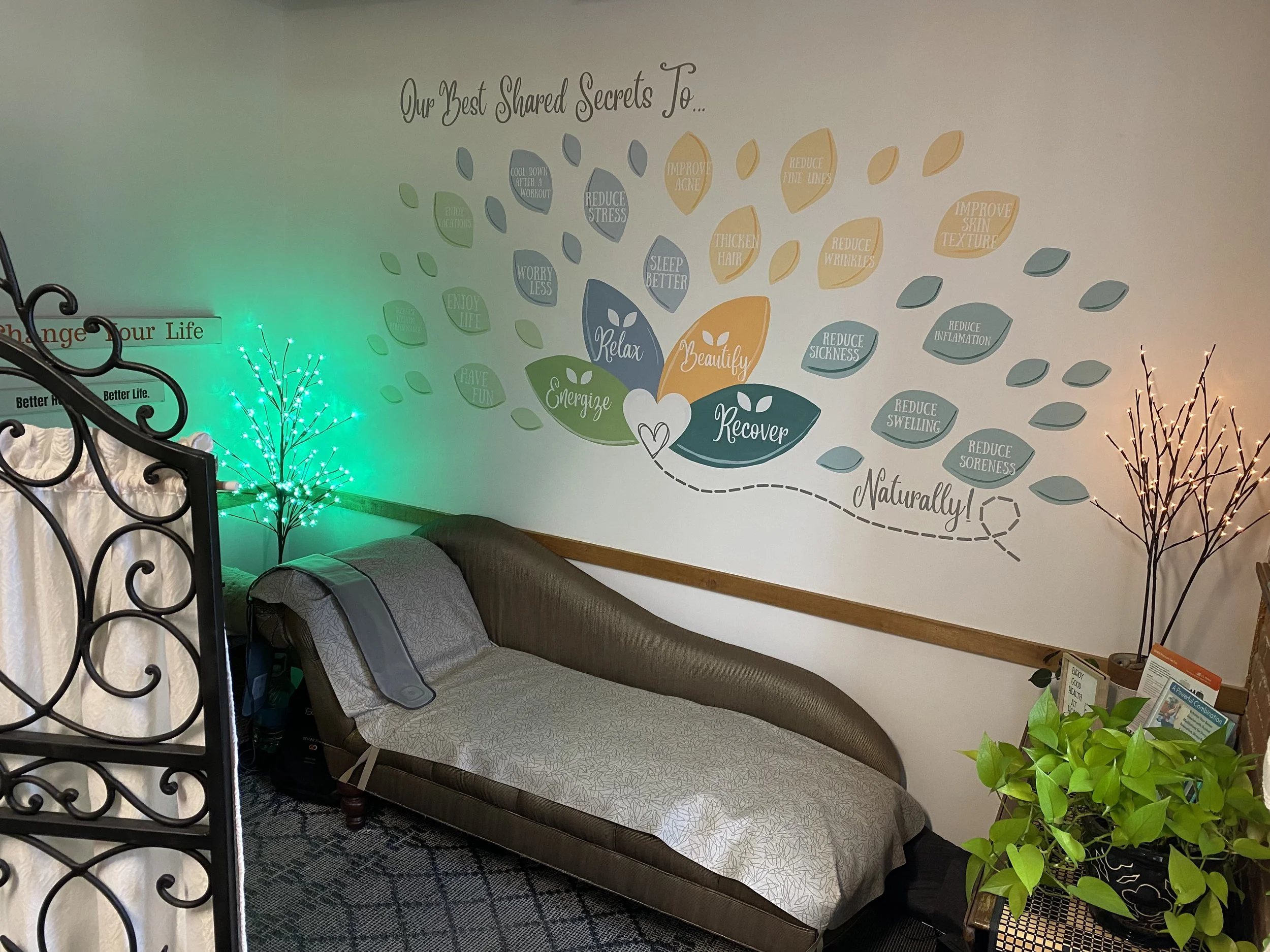 A cozy room with a lounge chair, indoor plants, and a wall decorated with a colorful mural that says "Our Best Shared Secrets To..." followed by various wellness and self-care benefits organized in petal shapes, with words like "Relax," "Recover," "Energize," and "Beautify."