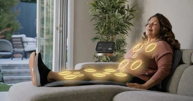A woman relaxing on a couch with glowing heated pads on her shoulders, stomach, and legs, with a control device nearby.