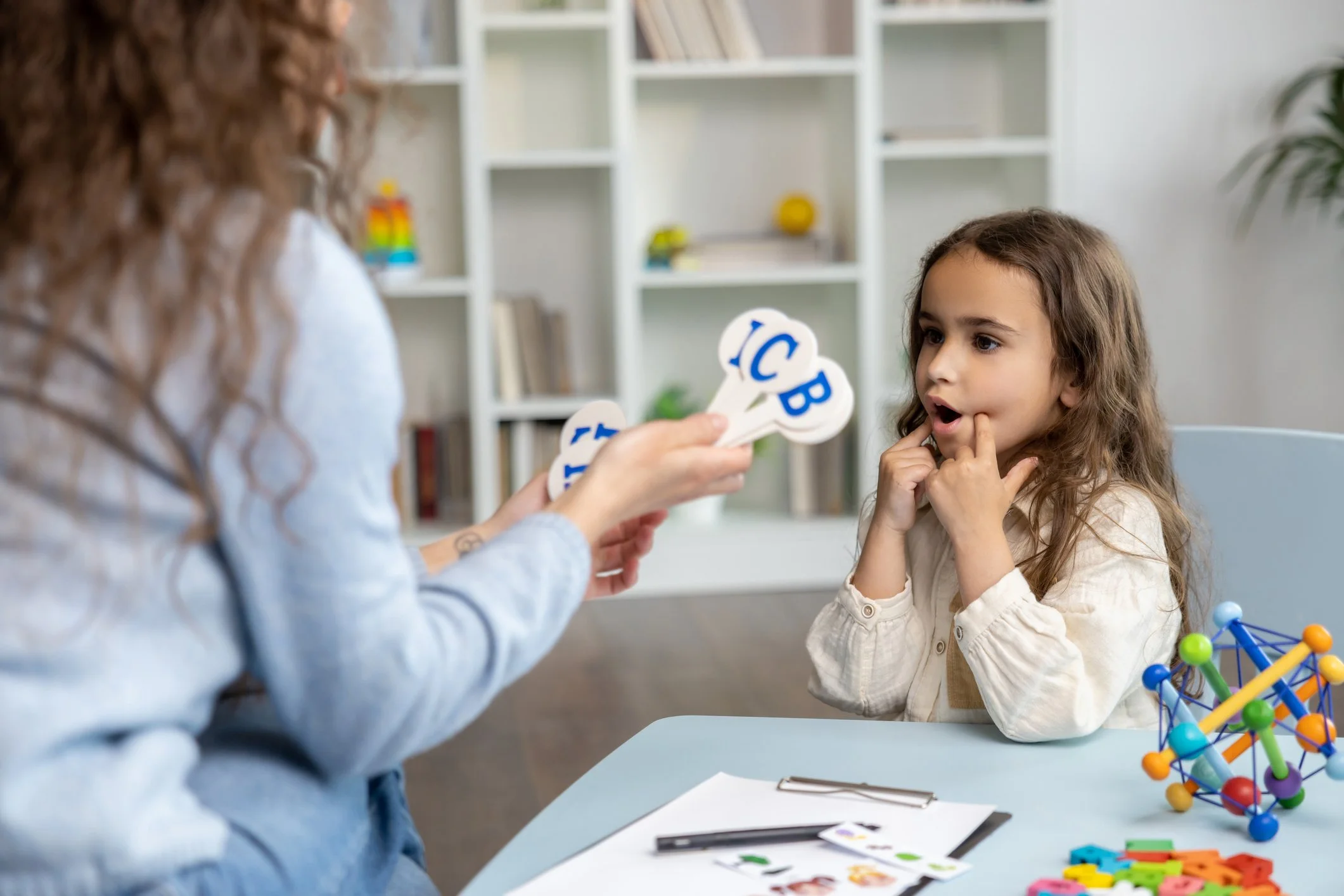 A young girl is sitting at a table with her fingers on her lips, looking at an adult who is holding flashcards with the alphabet. The girl is engaged in a speech therapy activity with educational toys and materials on the table.
