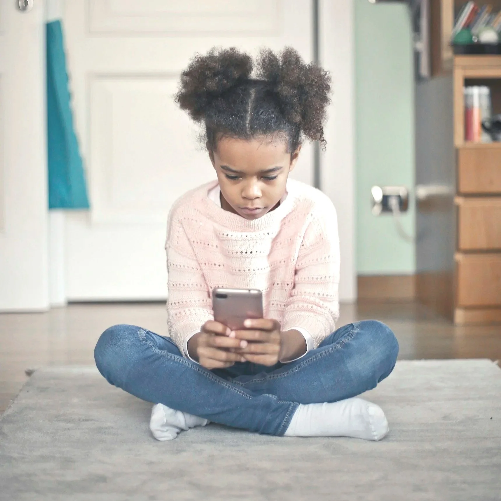 Young girl sitting cross-legged on a carpet, looking at a smartphone in her hands, in a room with a door and bookshelves in the background.
