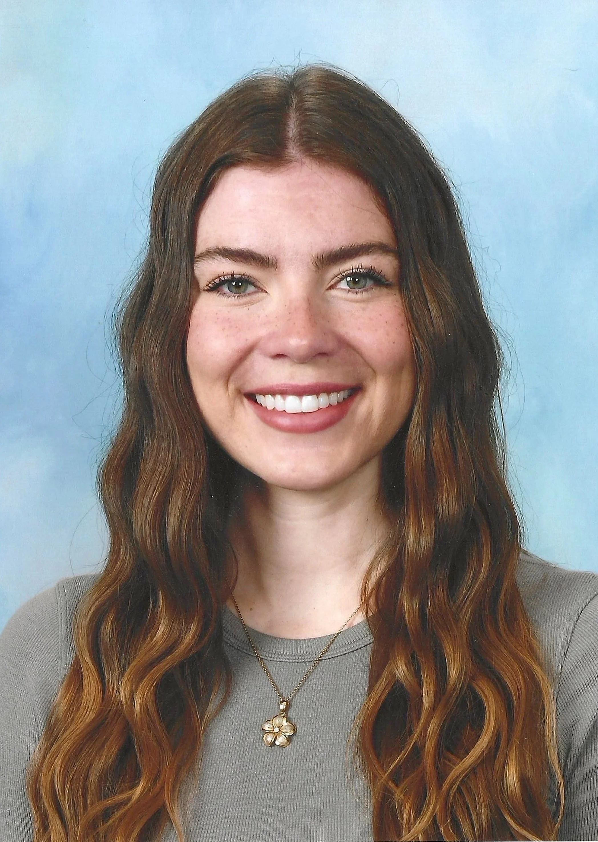 A young woman with long, wavy brown hair, blue eyes, and freckles, smiling in front of a light blue background, wearing a gray top and a gold flower-shaped pendant necklace.