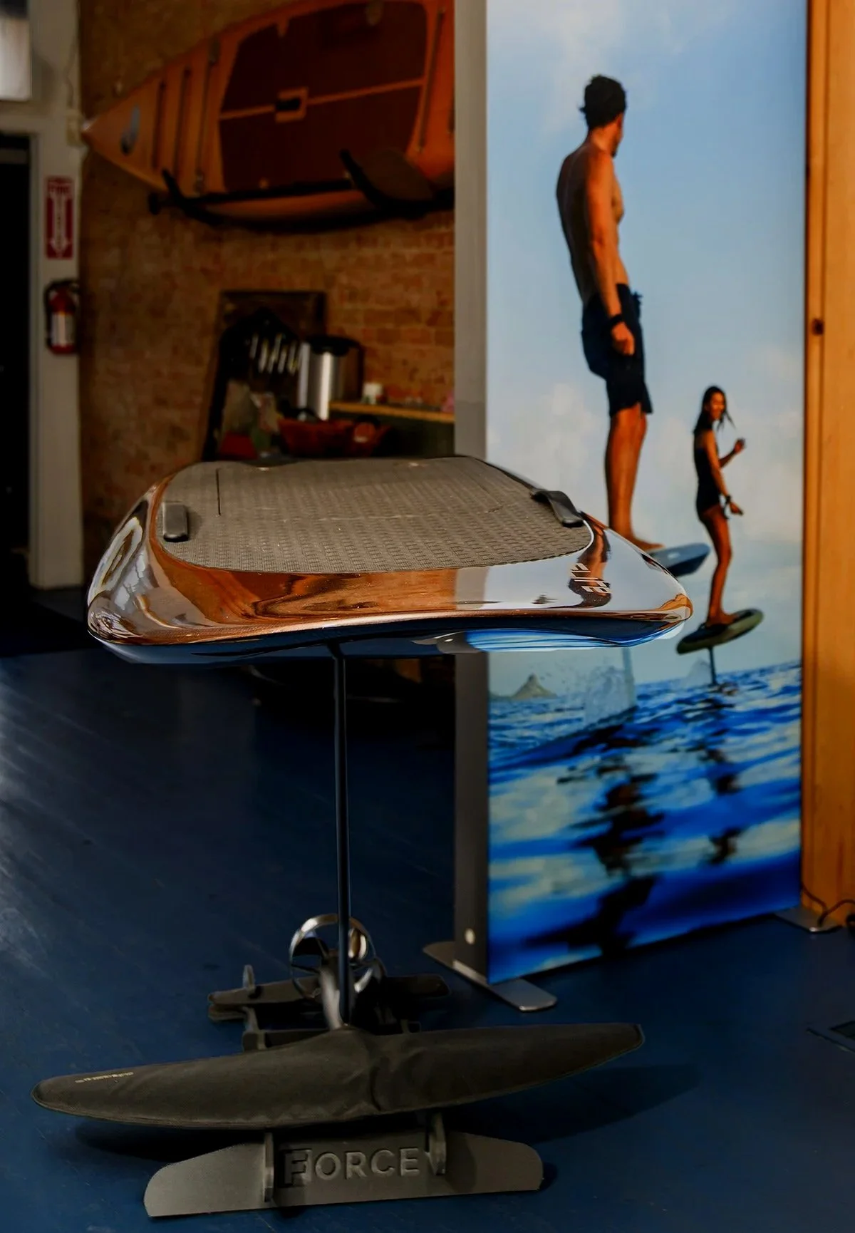 Indoor scene with a balance board stand and a mini surfboard in front, against a colorful mural of two people surfing on the water, with a brick wall, a paddleboard on the wall, and a fire extinguisher visible in the background.
