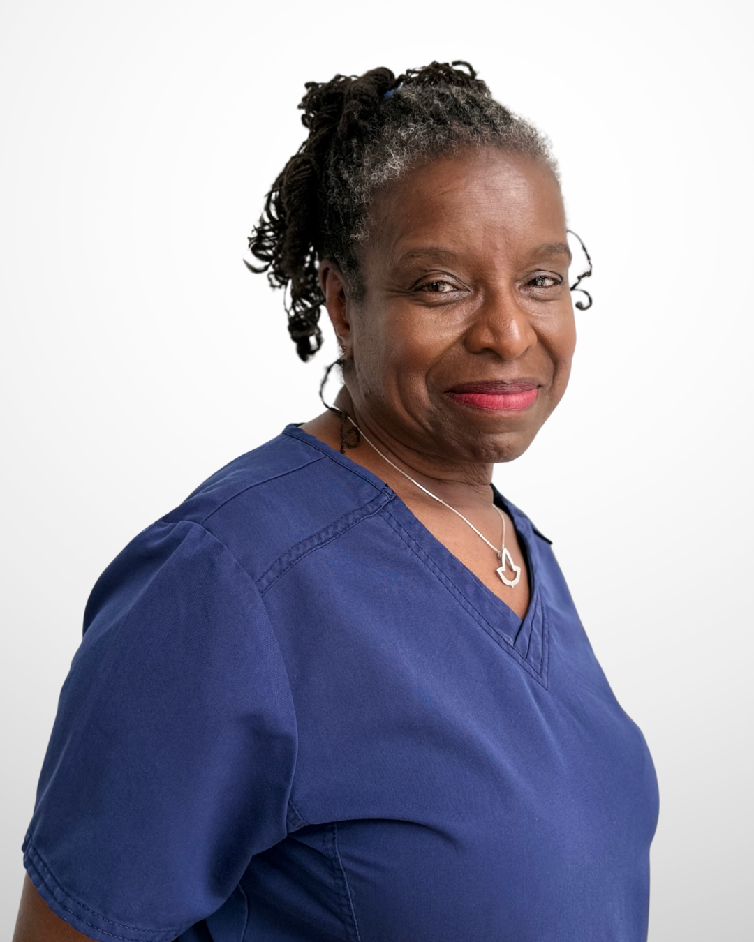 A mature African American woman with short, curly gray hair wearing blue medical scrubs and a silver necklace with a maple leaf pendant, smiling softly at the camera.