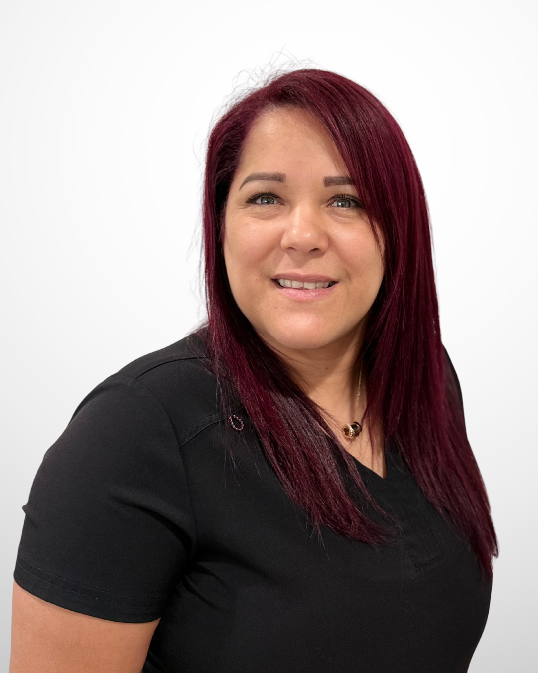 Portrait of a woman with long dark red hair, wearing a black top and a small necklace, standing against a plain white background.