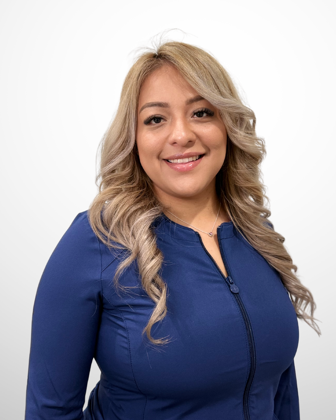A smiling woman with blonde wavy hair wearing a blue zip-up top against a plain white background.