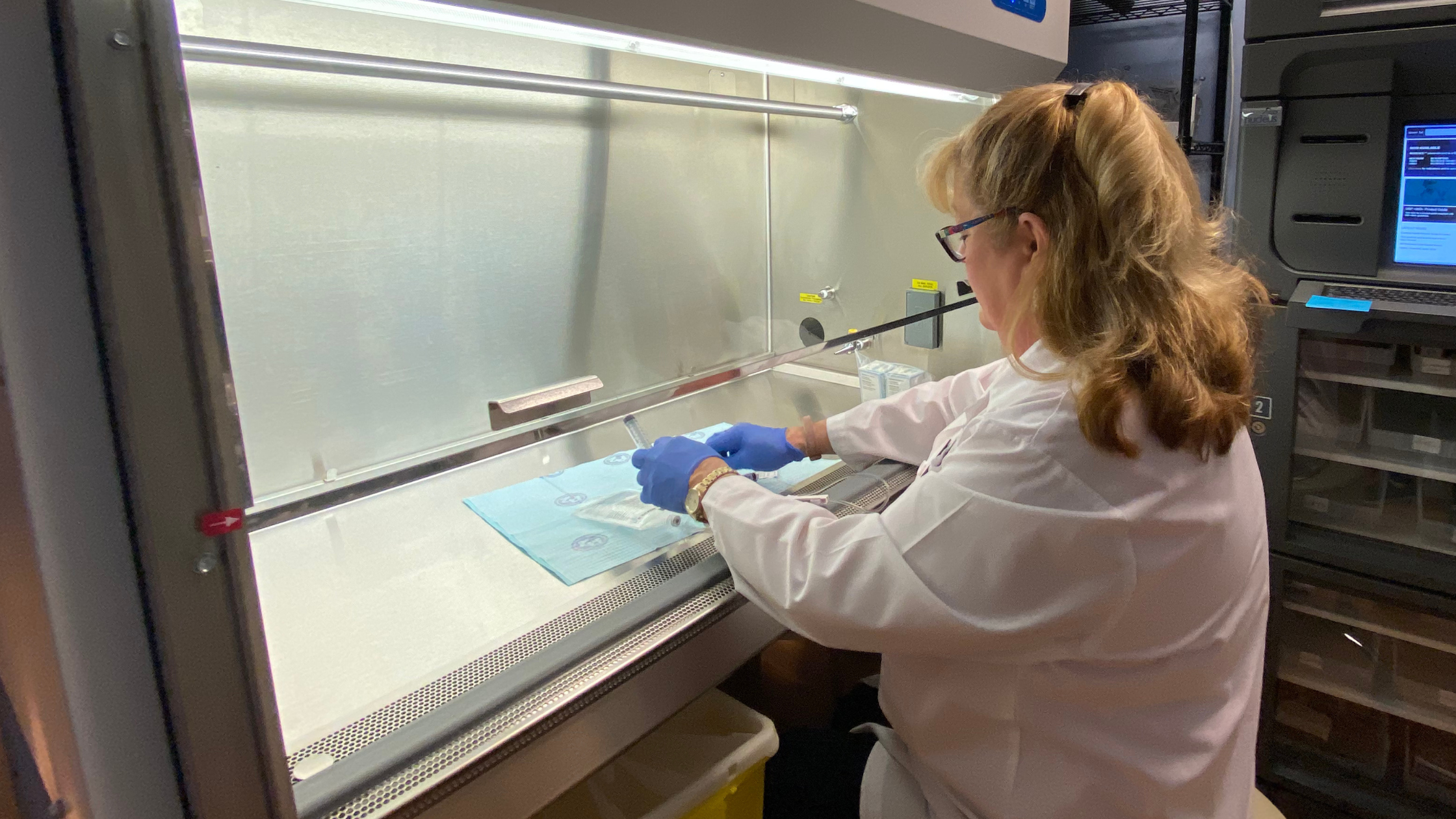 A scientist or healthcare worker working inside a laboratory fume hood, handling a sample with a pipette, wearing gloves, a white lab coat, and glasses.