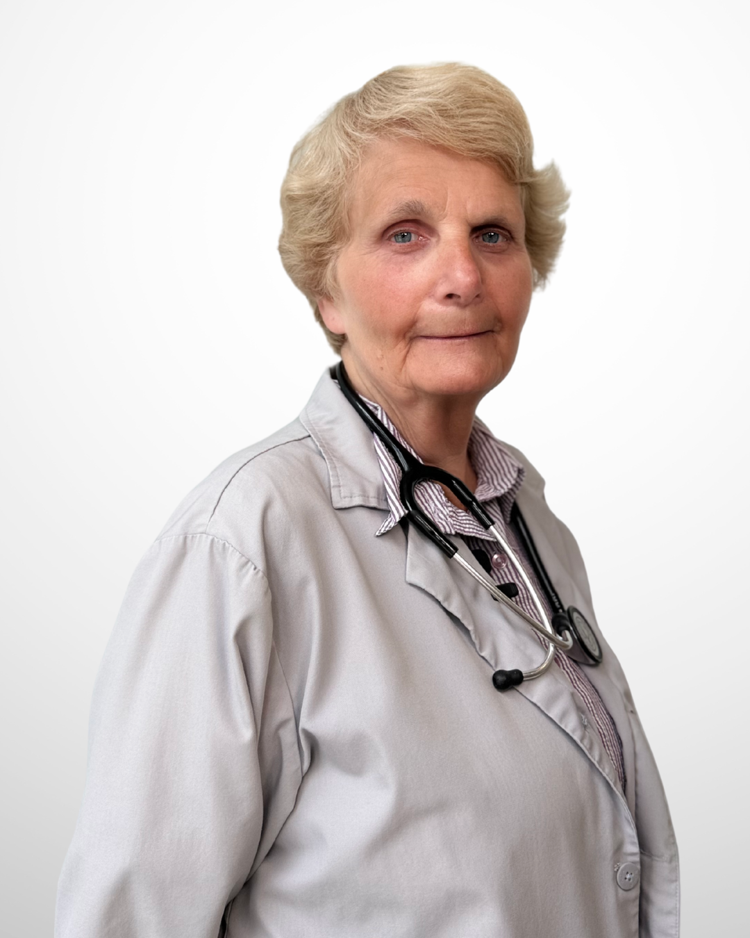 An elderly female doctor with short blonde hair wearing a light gray coat and a stethoscope around her neck, standing against a plain white background.