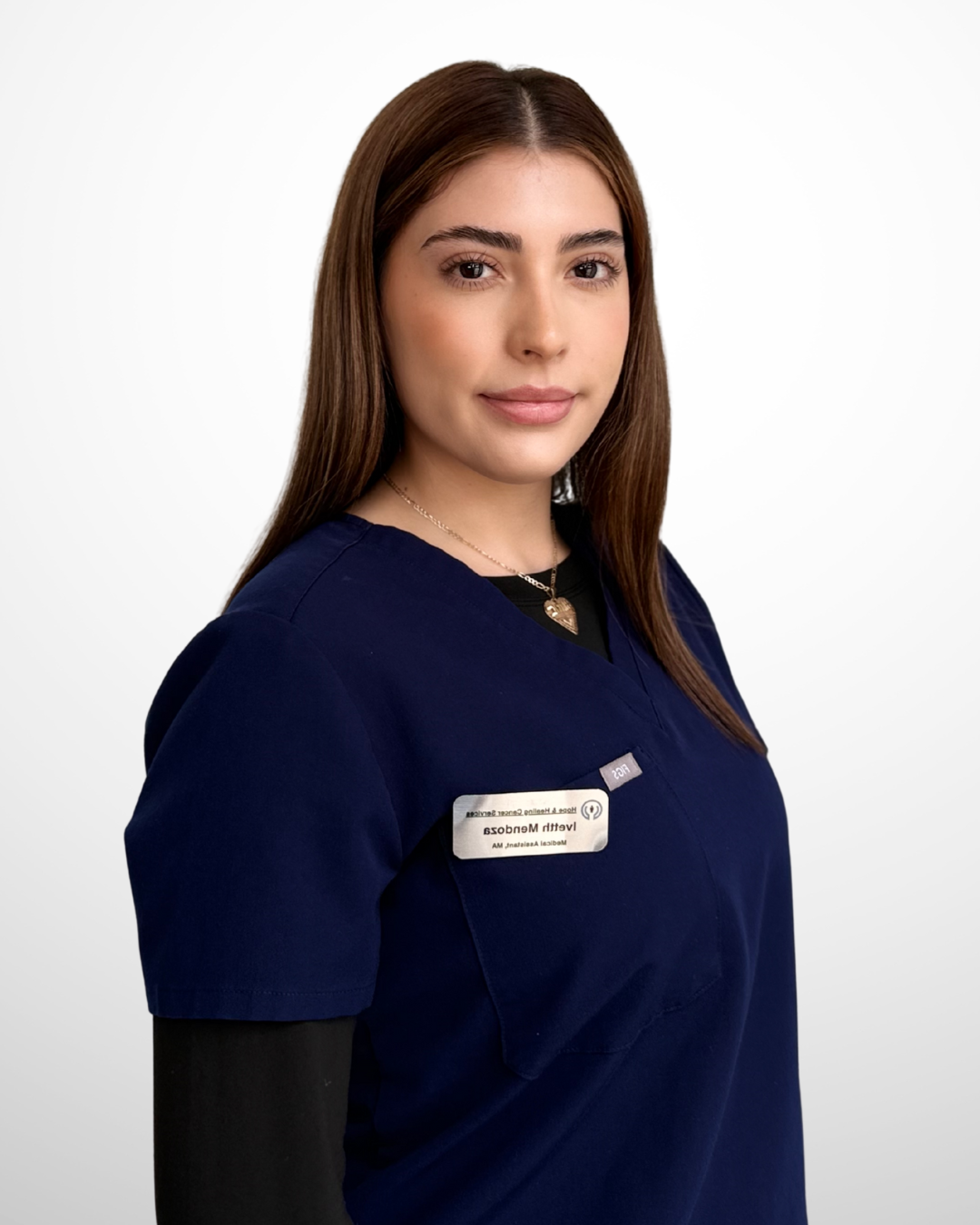 A young woman with long brown hair wearing navy blue medical scrubs and a name badge, standing against a plain white background.