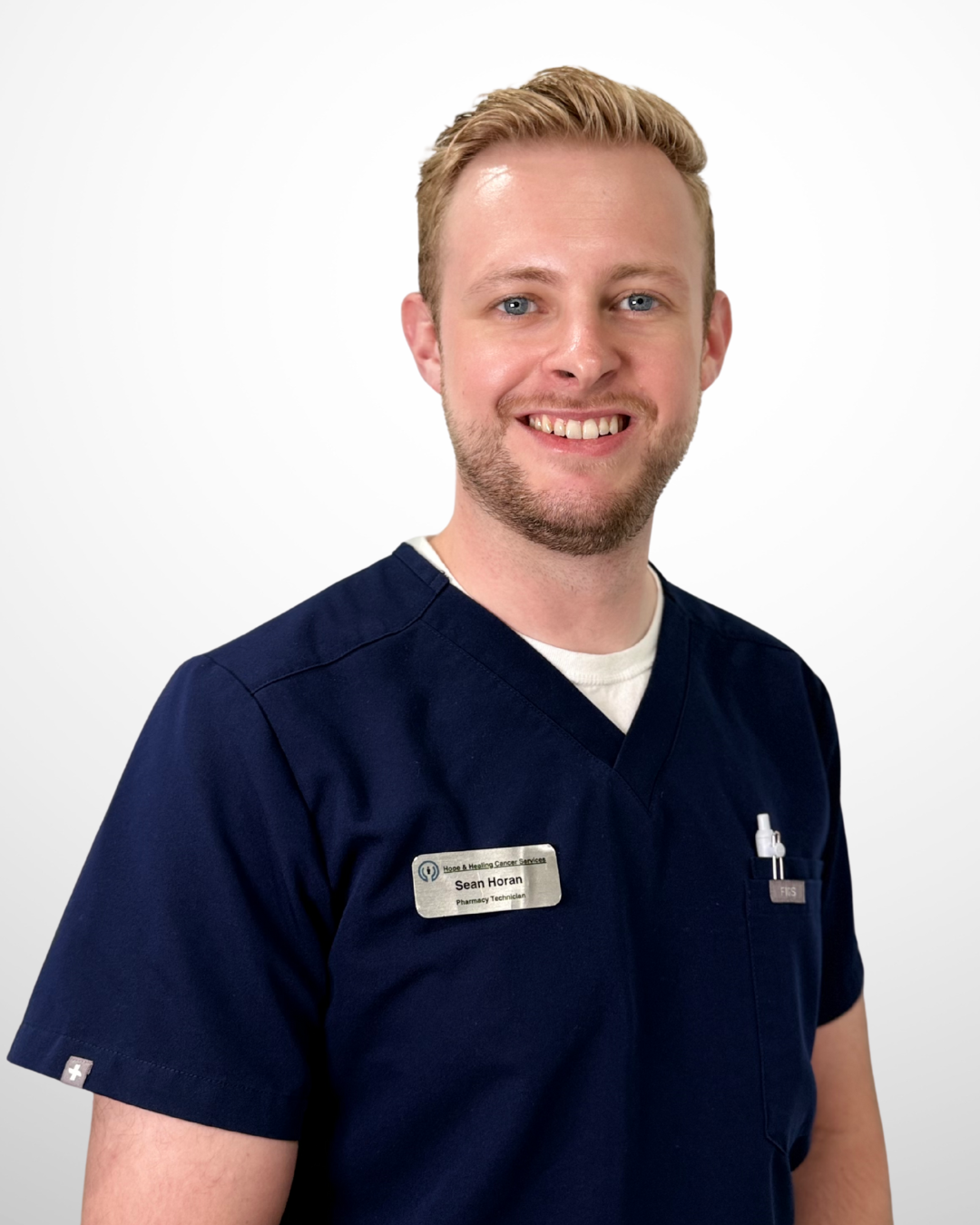 A smiling man with short blond hair and a beard, wearing navy medical scrubs and a name tag that reads 'Sean Horan, Pharmacy Technician.'