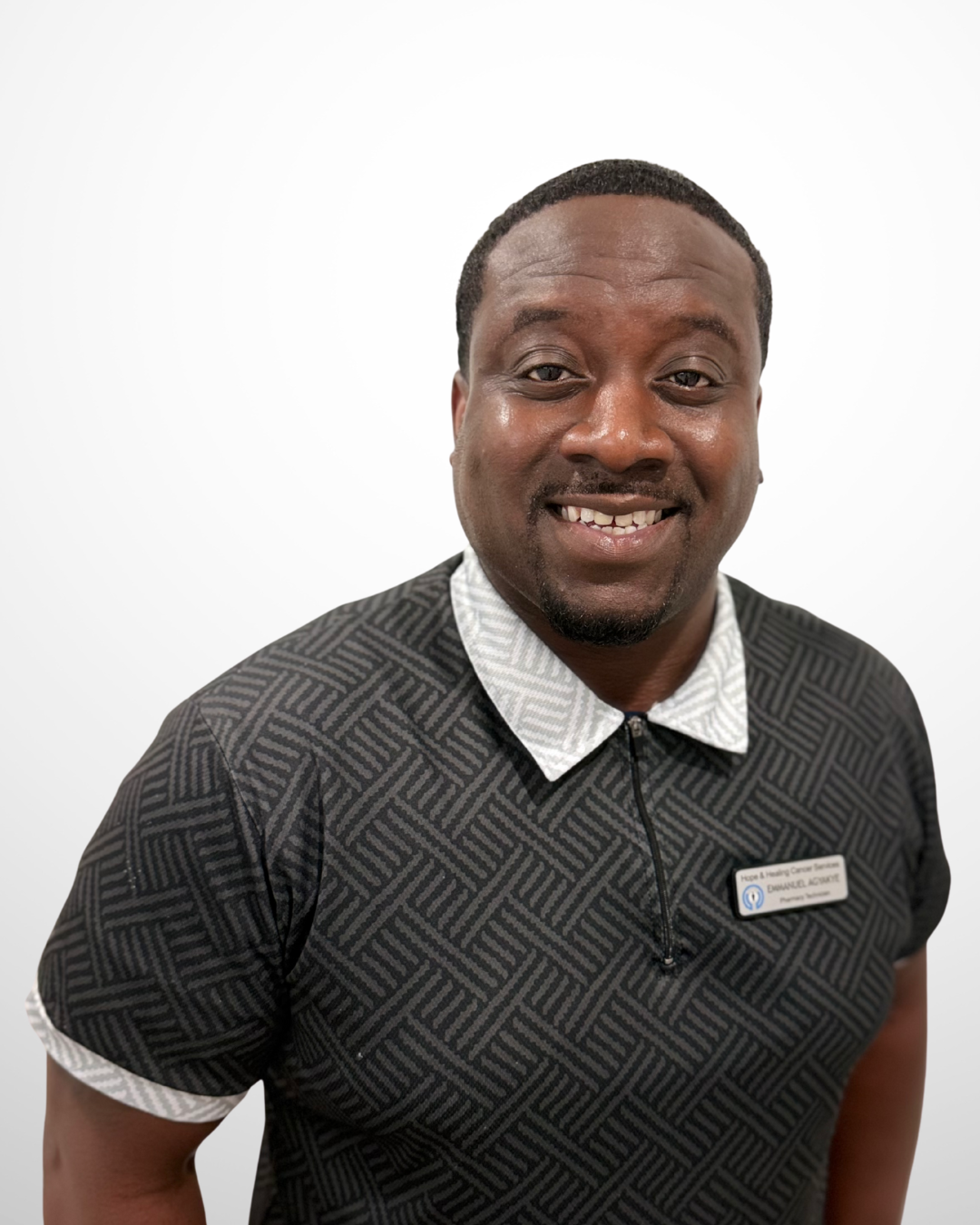 Portrait of a smiling man wearing a black patterned shirt with a name tag against a white background.