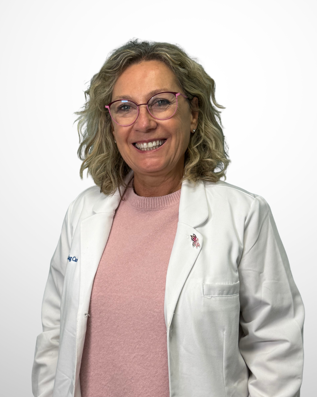 A woman with curly blonde hair, wearing pink glasses, a white medical coat, and a pink sweater smiling at the camera against a white background.