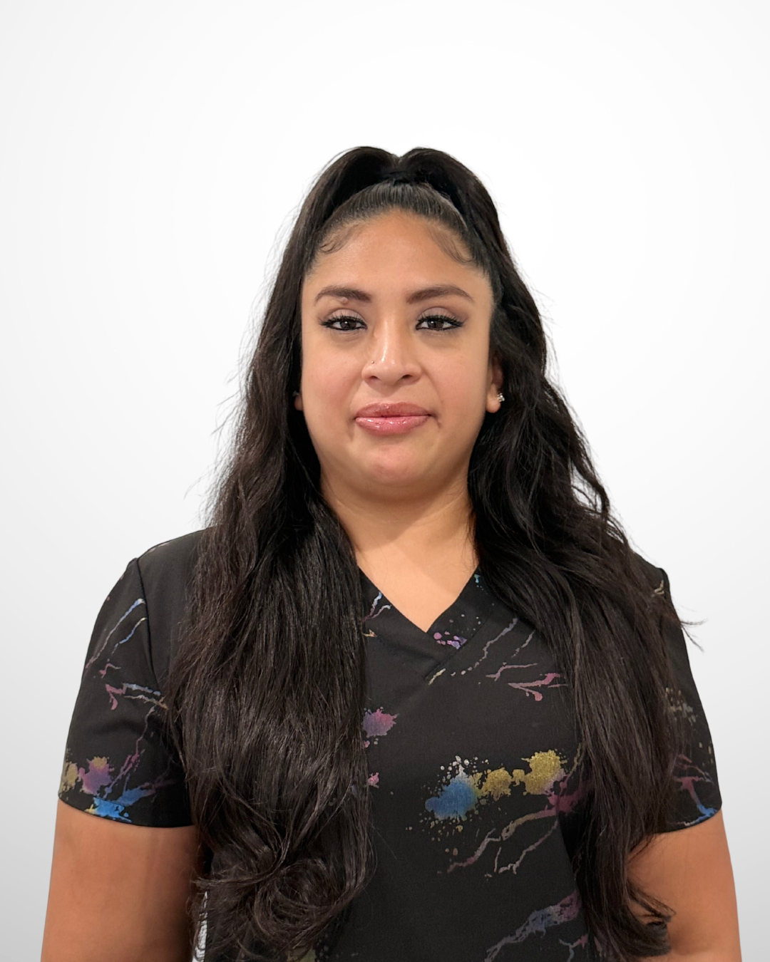 A woman with long, dark hair styled in a high half ponytail, wearing a black shirt with colorful paint splatter patterns, standing against a plain white background.