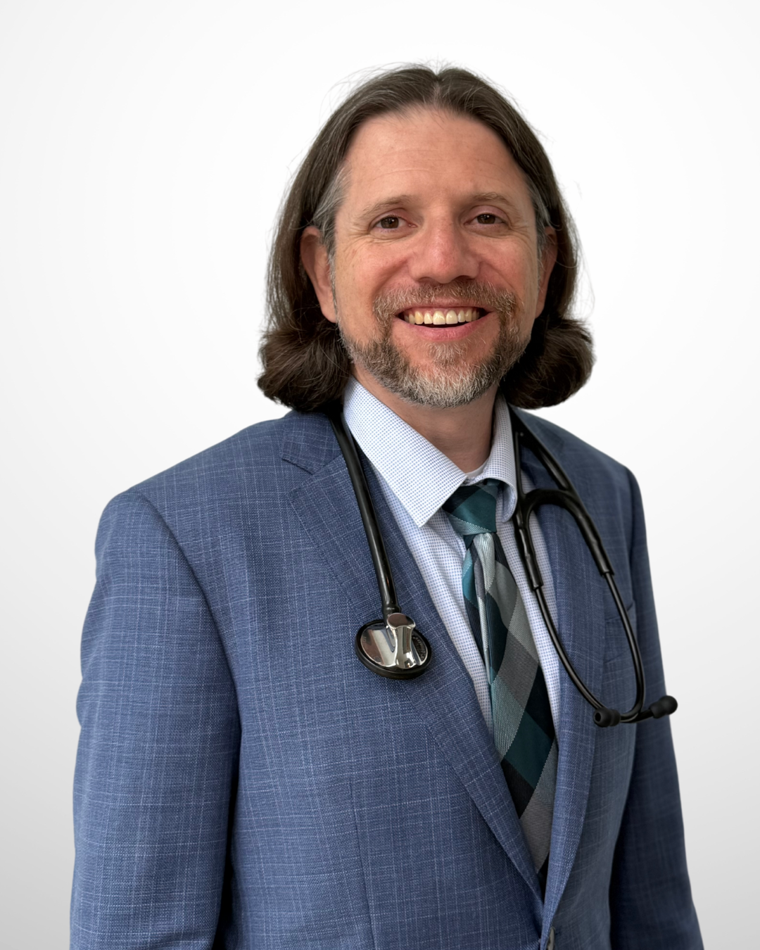 A smiling male doctor in a blue suit with a stethoscope around his neck, wearing a shirt and tie, against a plain white background.