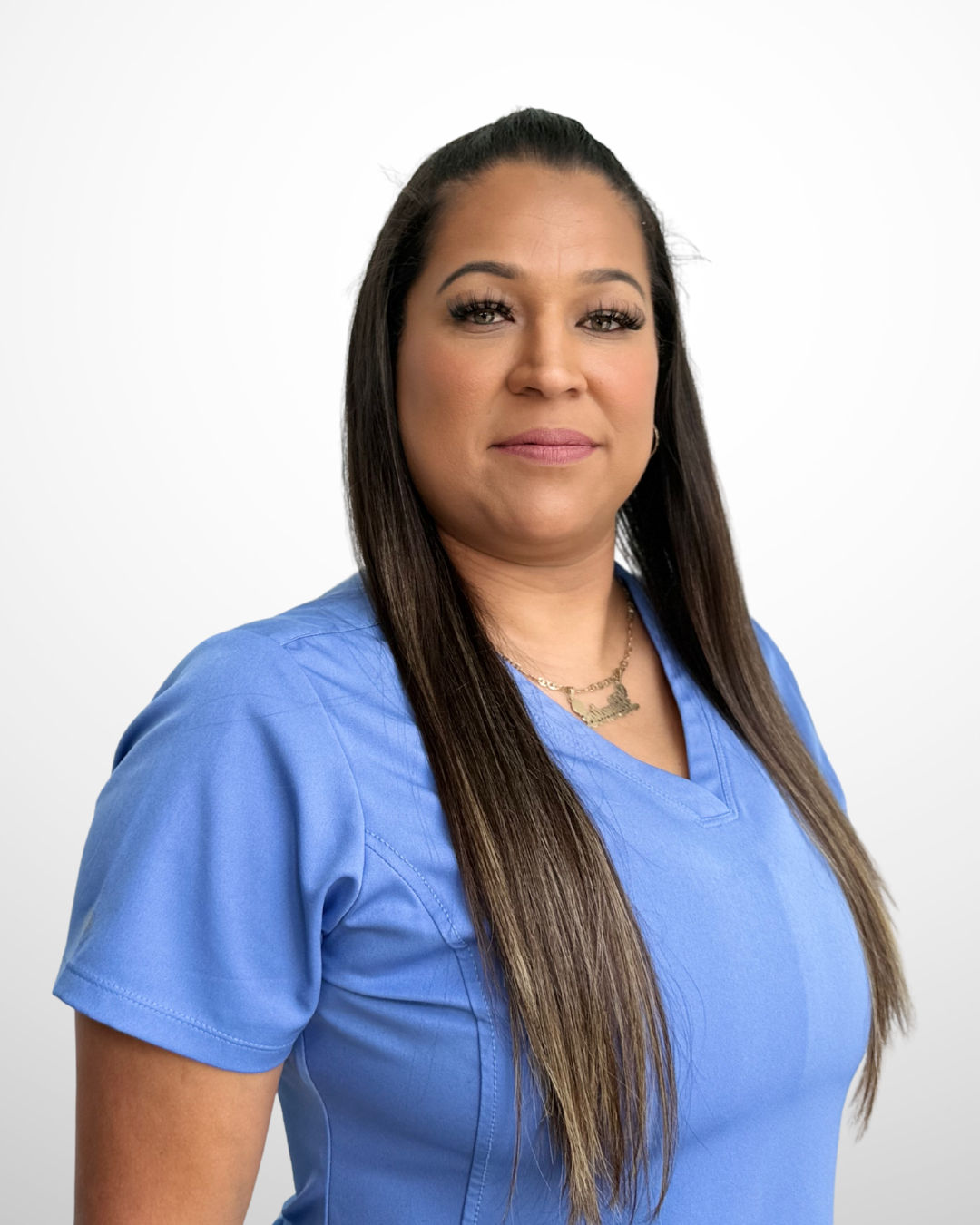 Portrait of a woman wearing blue scrubs, with long dark hair, looking at the camera against a white background.